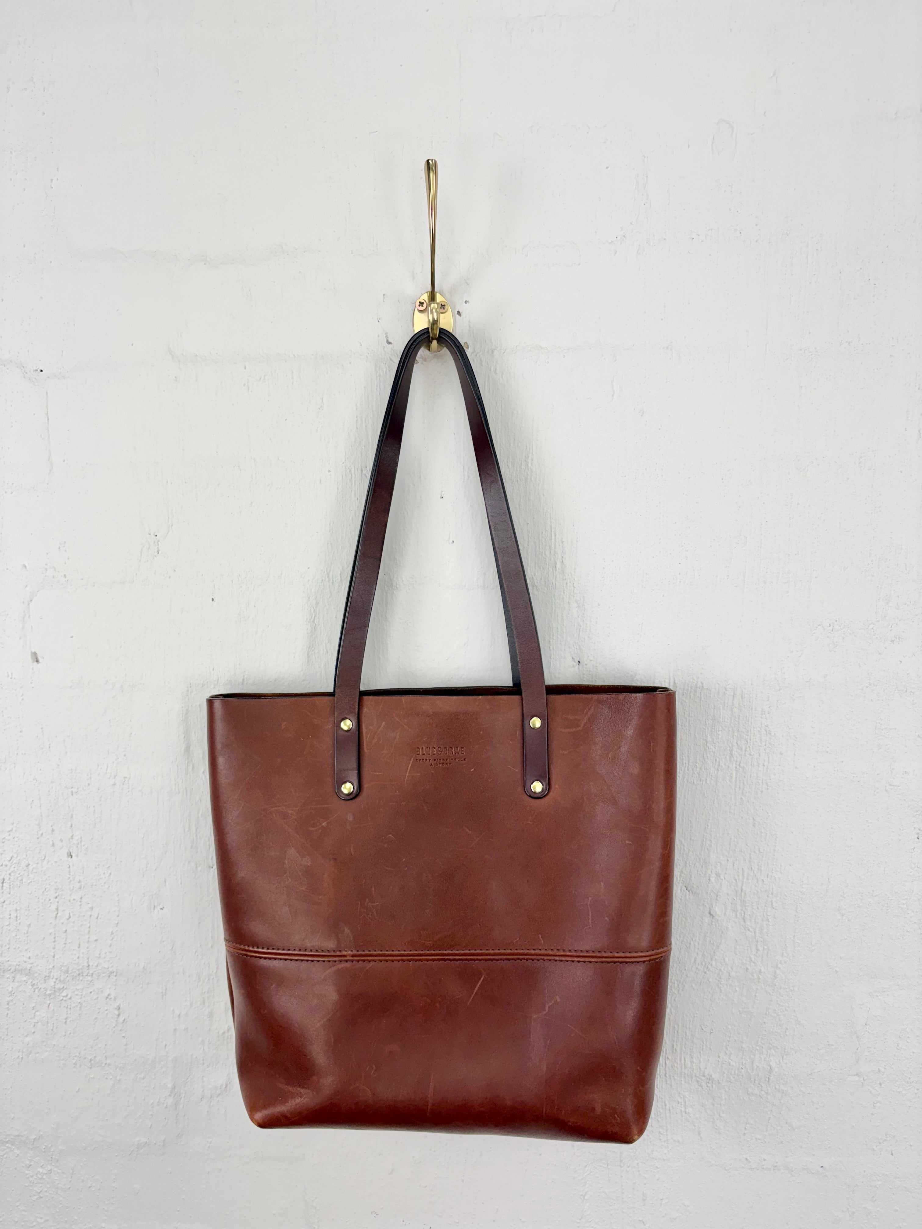 A brown leather tote bag with dark brown shoulder straps and gold-tone hardware, hanging from a gold hook against a white painted brick wall.