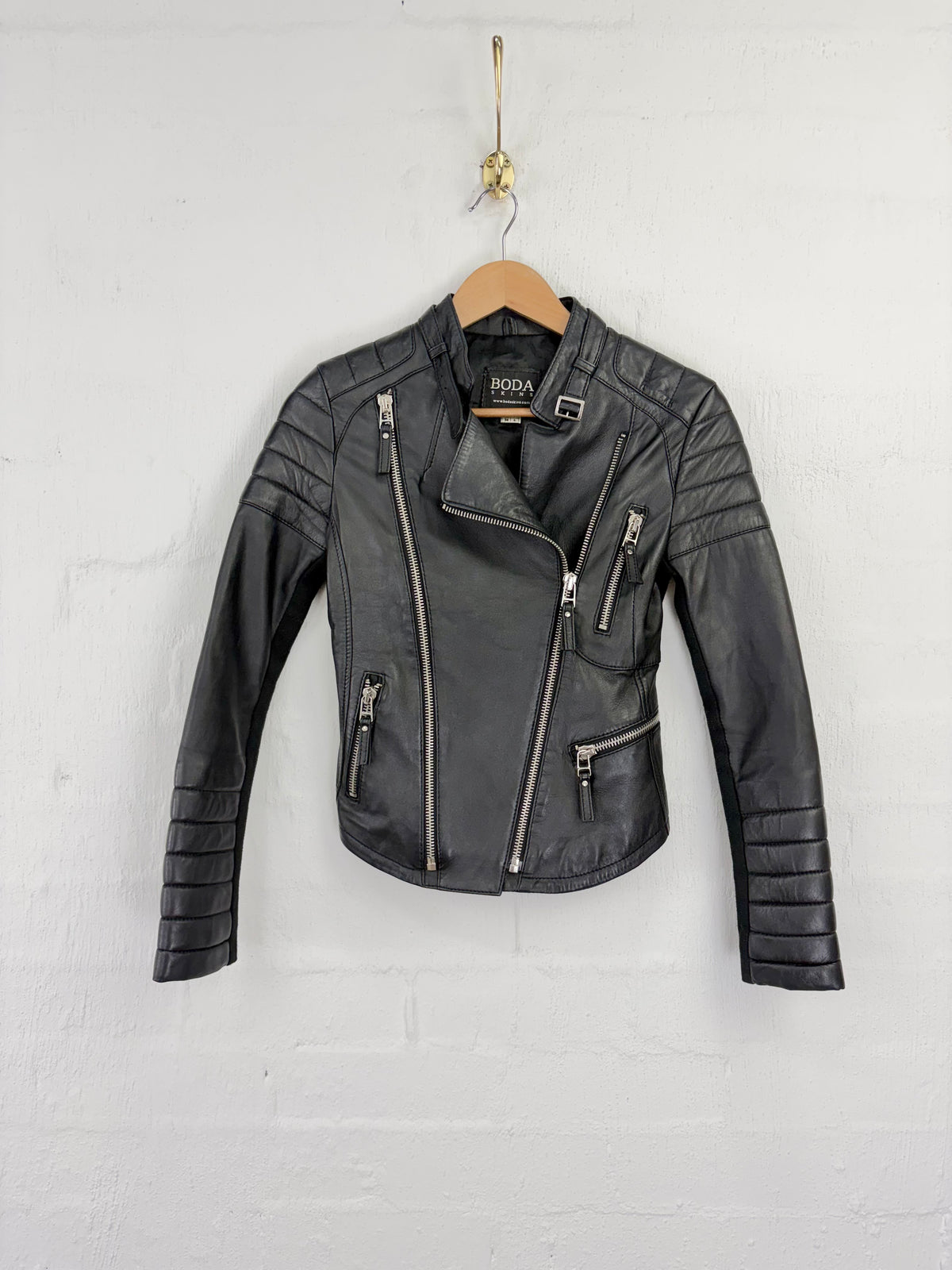 A black faux leather motorcycle jacket with silver zippers, hanging on a wooden hanger against a white painted brick wall.