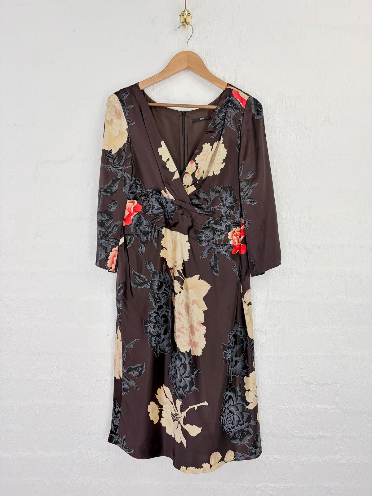 A dark brown wrap dress with a beige and black floral print and red flower accents, hanging on a wooden hanger against a white brick wall.