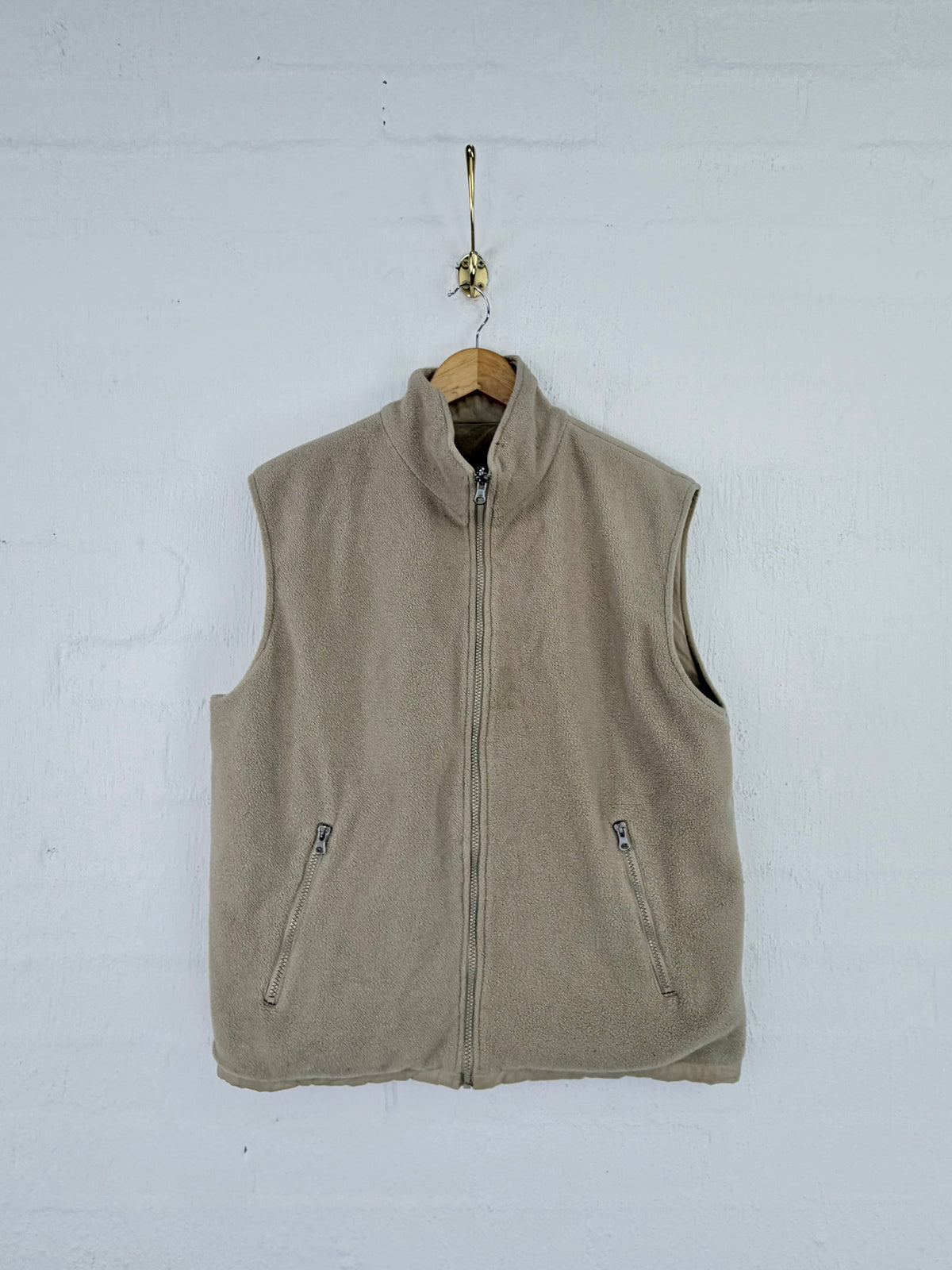 A beige fleece vest with a full zipper and zippered hand pockets, hanging on a wooden hanger against a white painted brick wall.