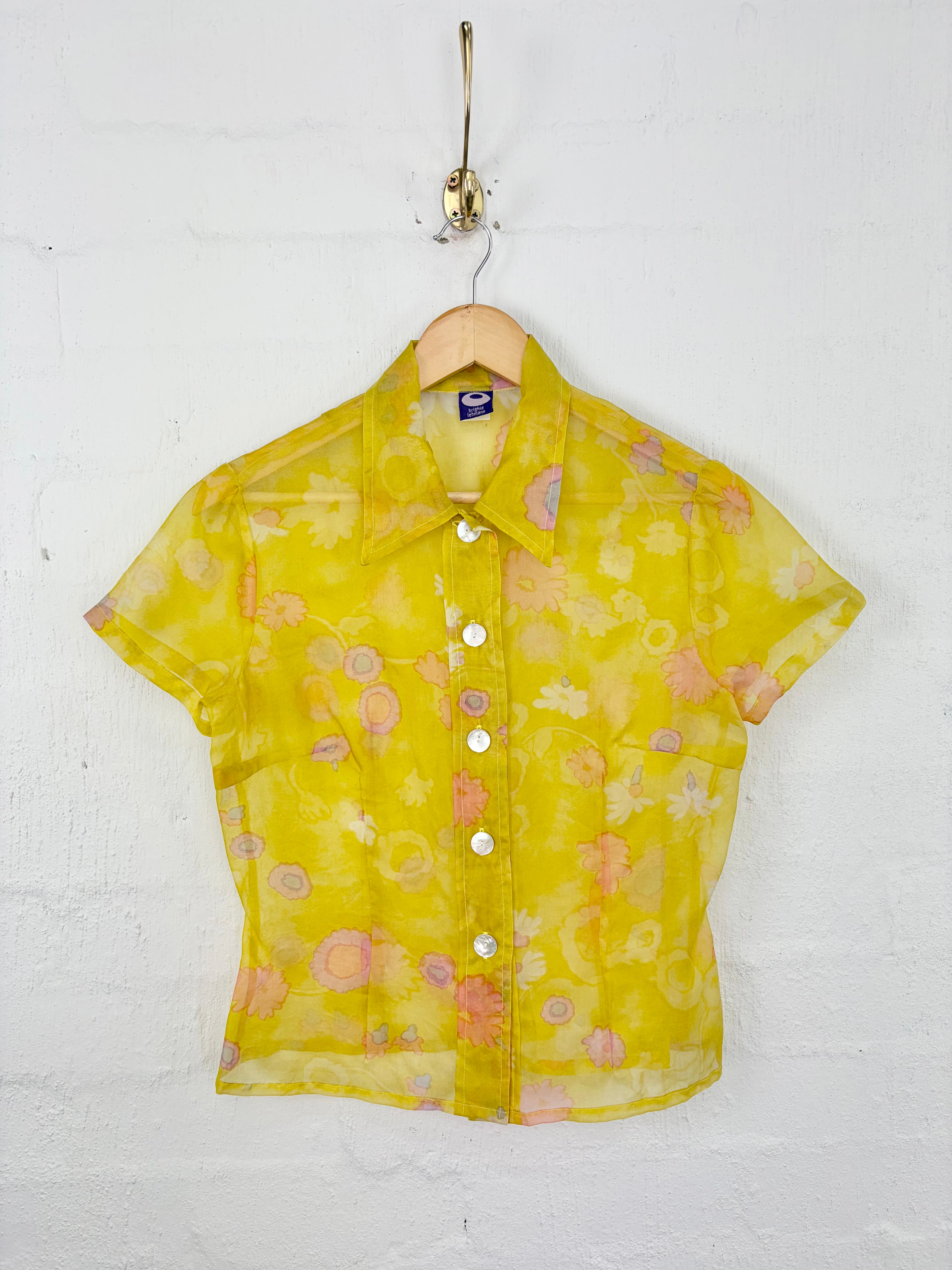 A yellow short-sleeve button-up shirt with a pink and orange floral pattern, hanging on a wooden hanger from a gold hook against a white brick wall.