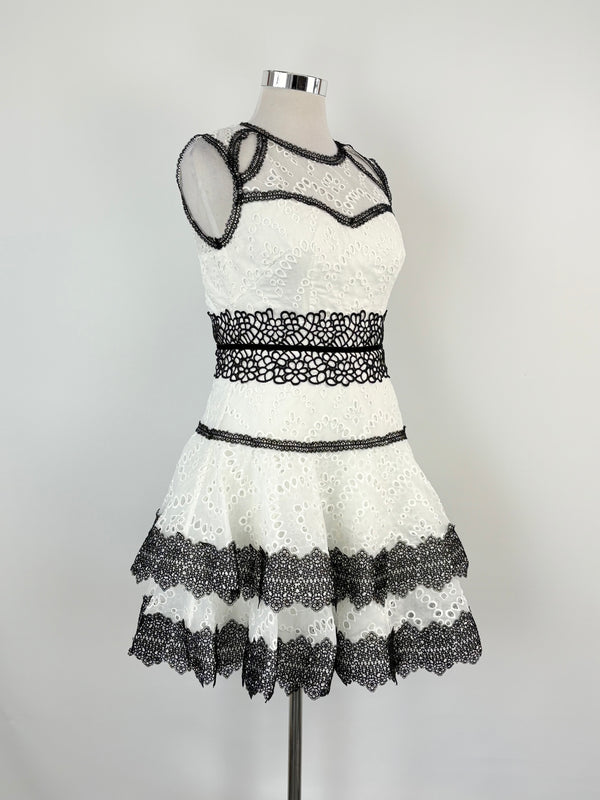 A white mini dress with black trim and embroidered details, displayed on a mannequin against a plain background.
