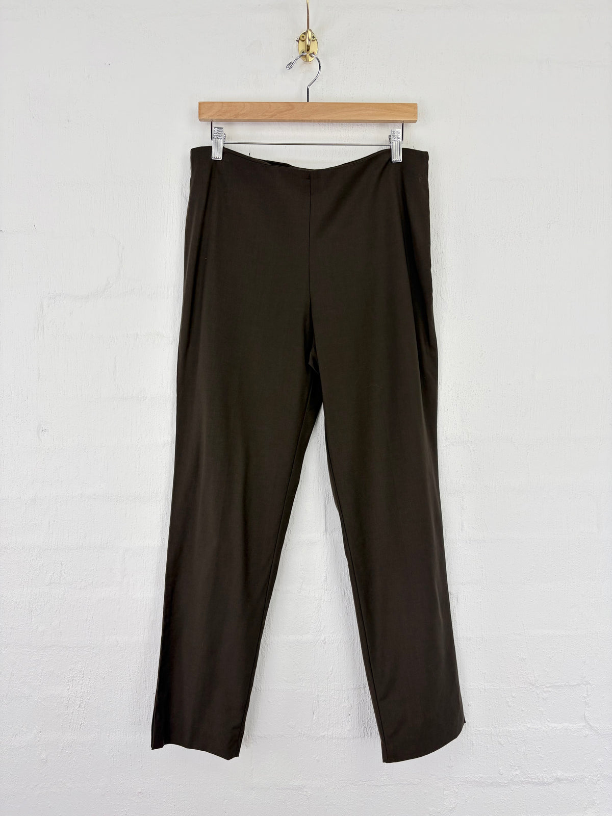 Dark brown wide-leg cropped pants hanging from a wooden hanger against a white painted brick wall.