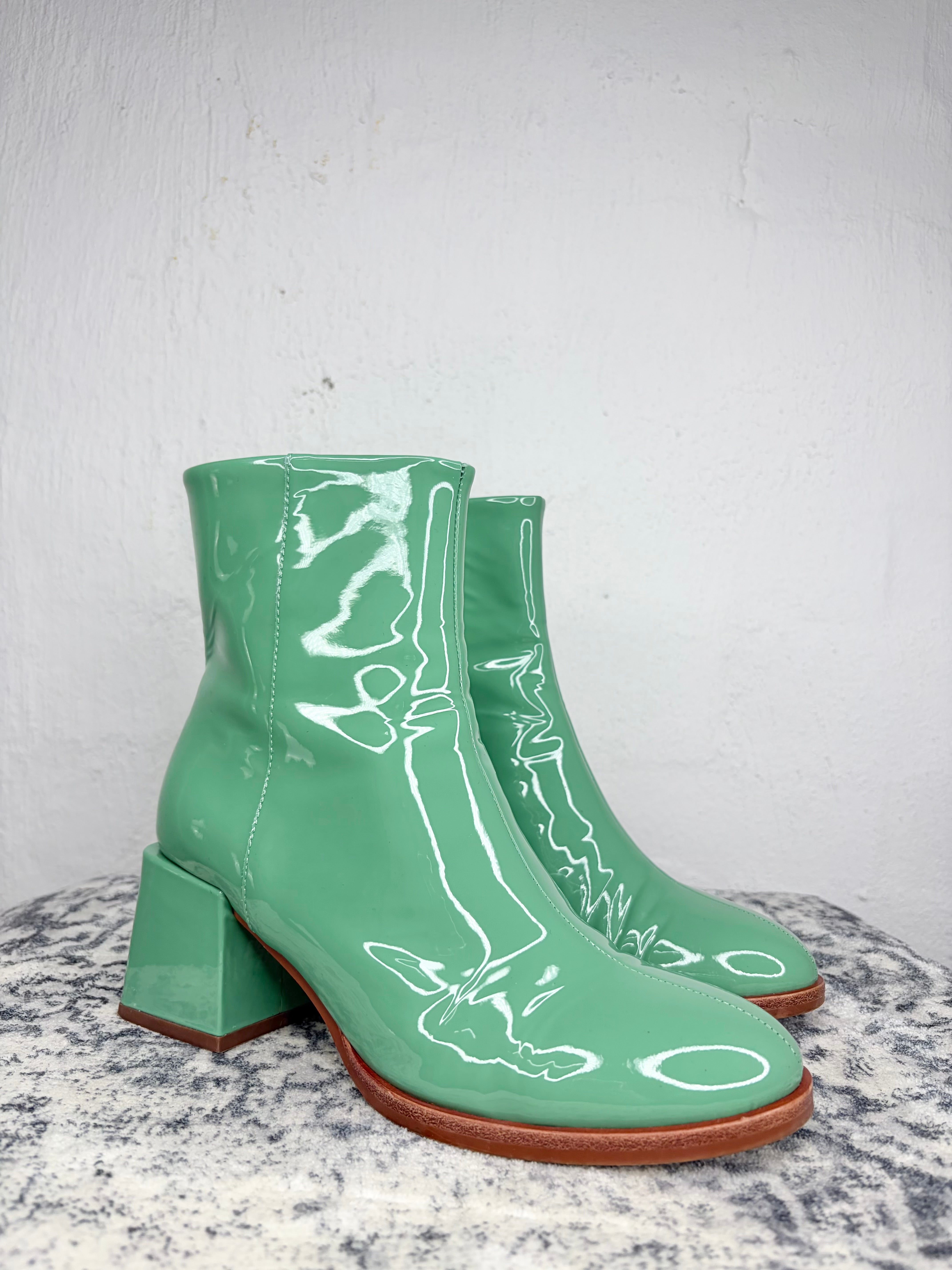 A pair of mint green patent leather ankle boots with block heels, displayed on a speckled gray stone surface against a white textured wall.
