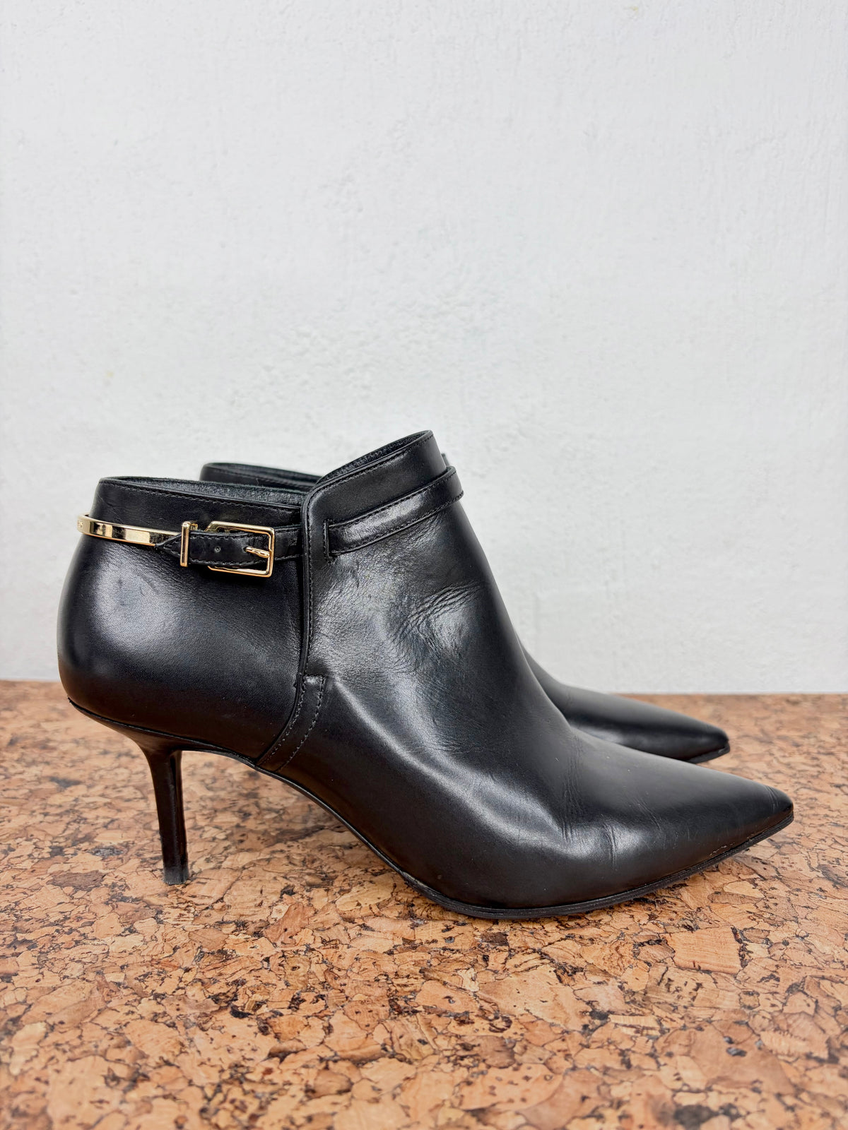 A pair of black leather ankle boots with pointed toes and slim kitten heels, featuring a decorative gold-tone buckle strap on the outer side, displayed on a cork surface against a light gray wall.