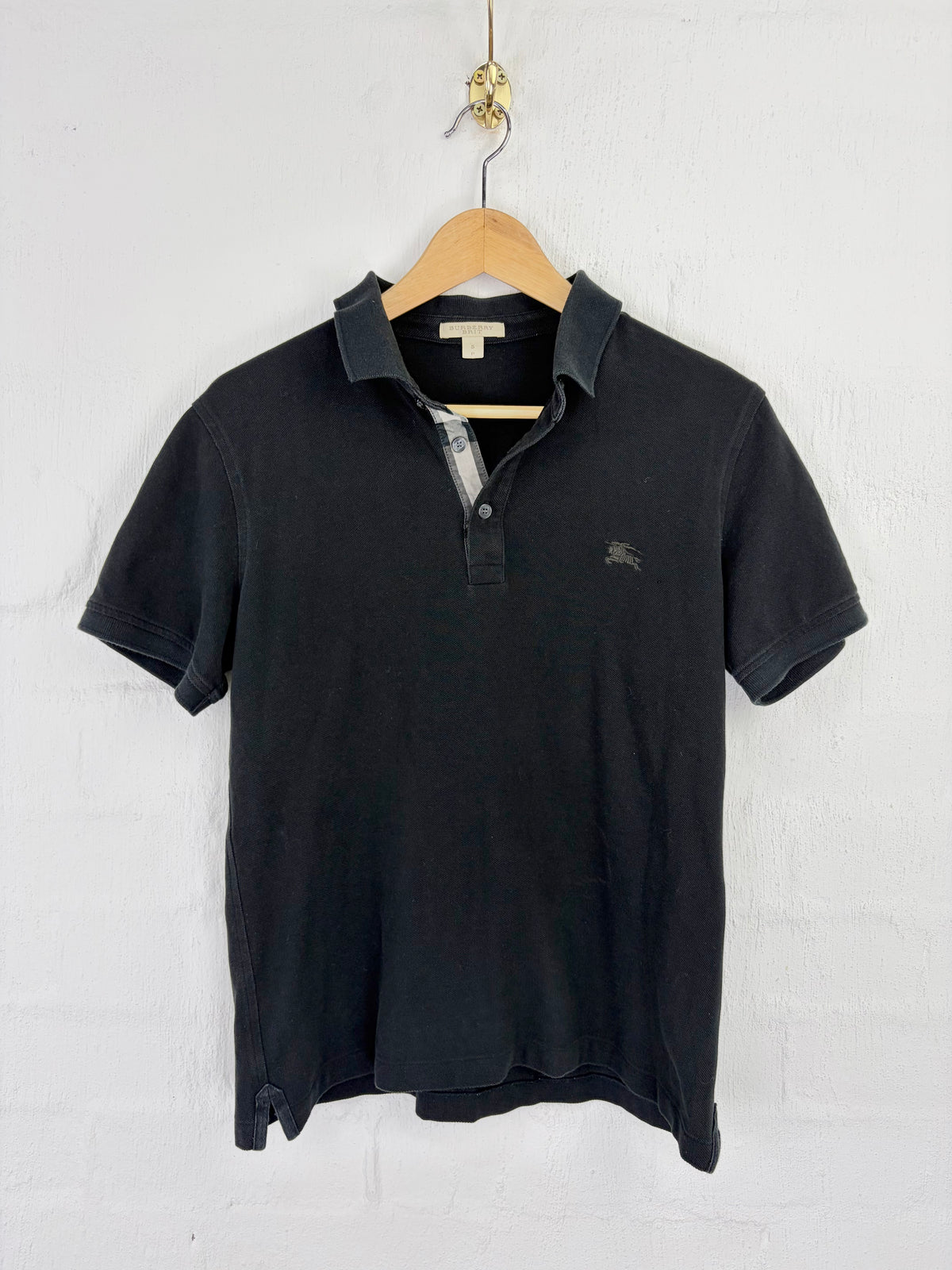 A black short-sleeve polo shirt hanging on a wooden hanger against a white painted brick wall. The shirt features a small embroidered horse logo on the left chest, indicating the brand Burberry.