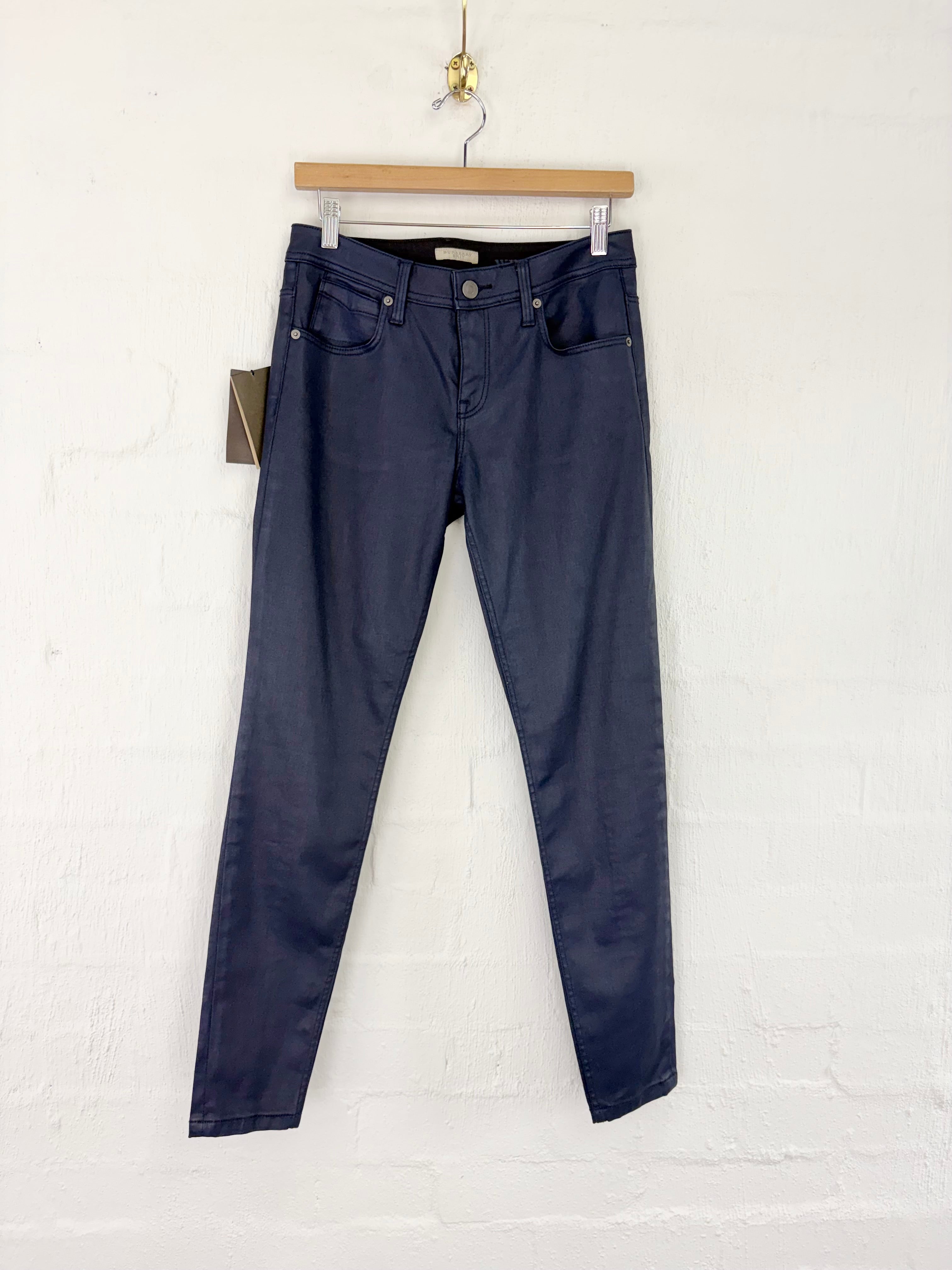 A pair of dark navy blue slim-fit jeans hanging from a wooden hanger against a white painted brick wall.
