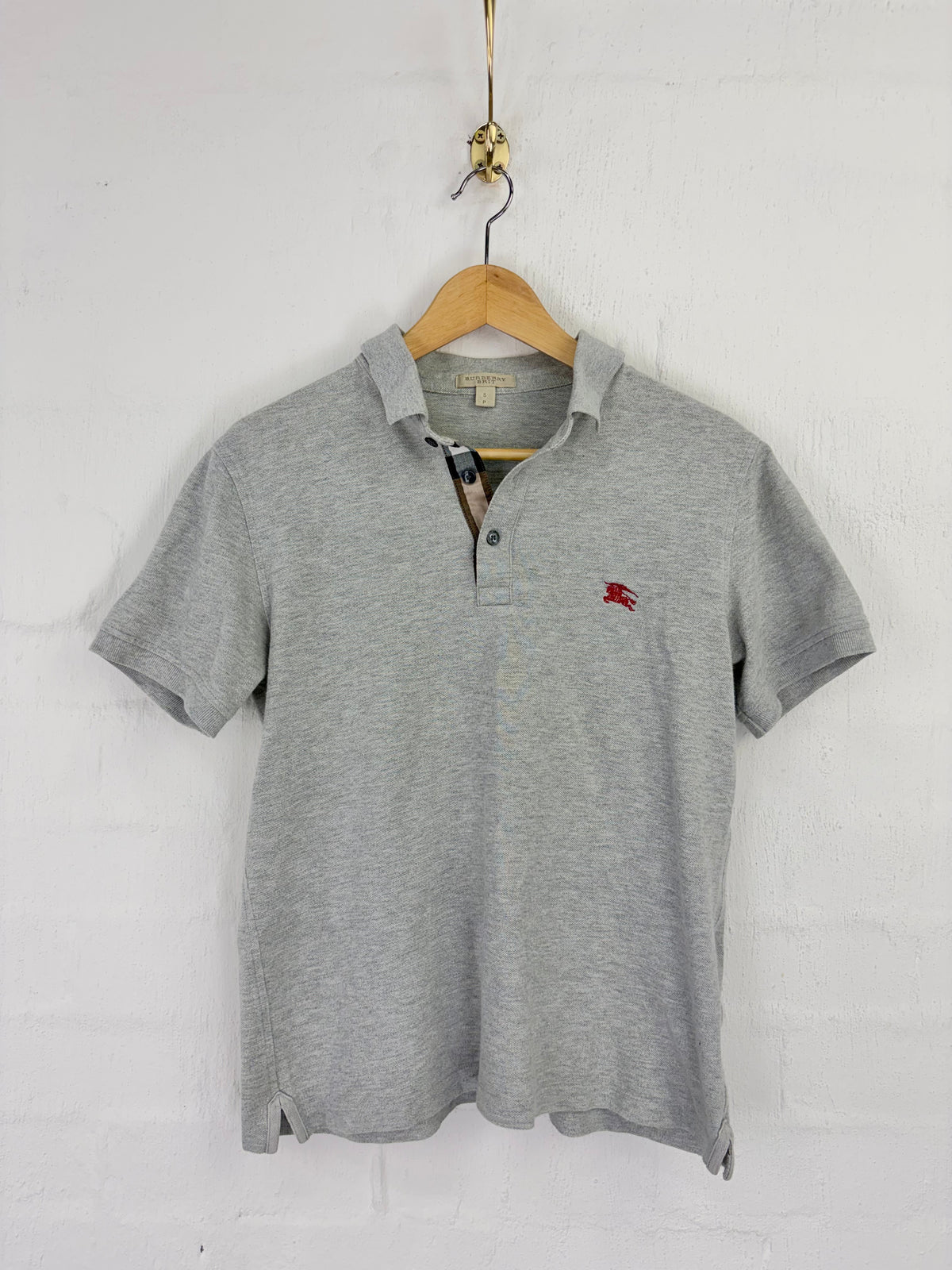 A heather gray short-sleeve polo shirt with a small red embroidered logo on the left chest, hanging on a wooden hanger against a white painted brick wall.