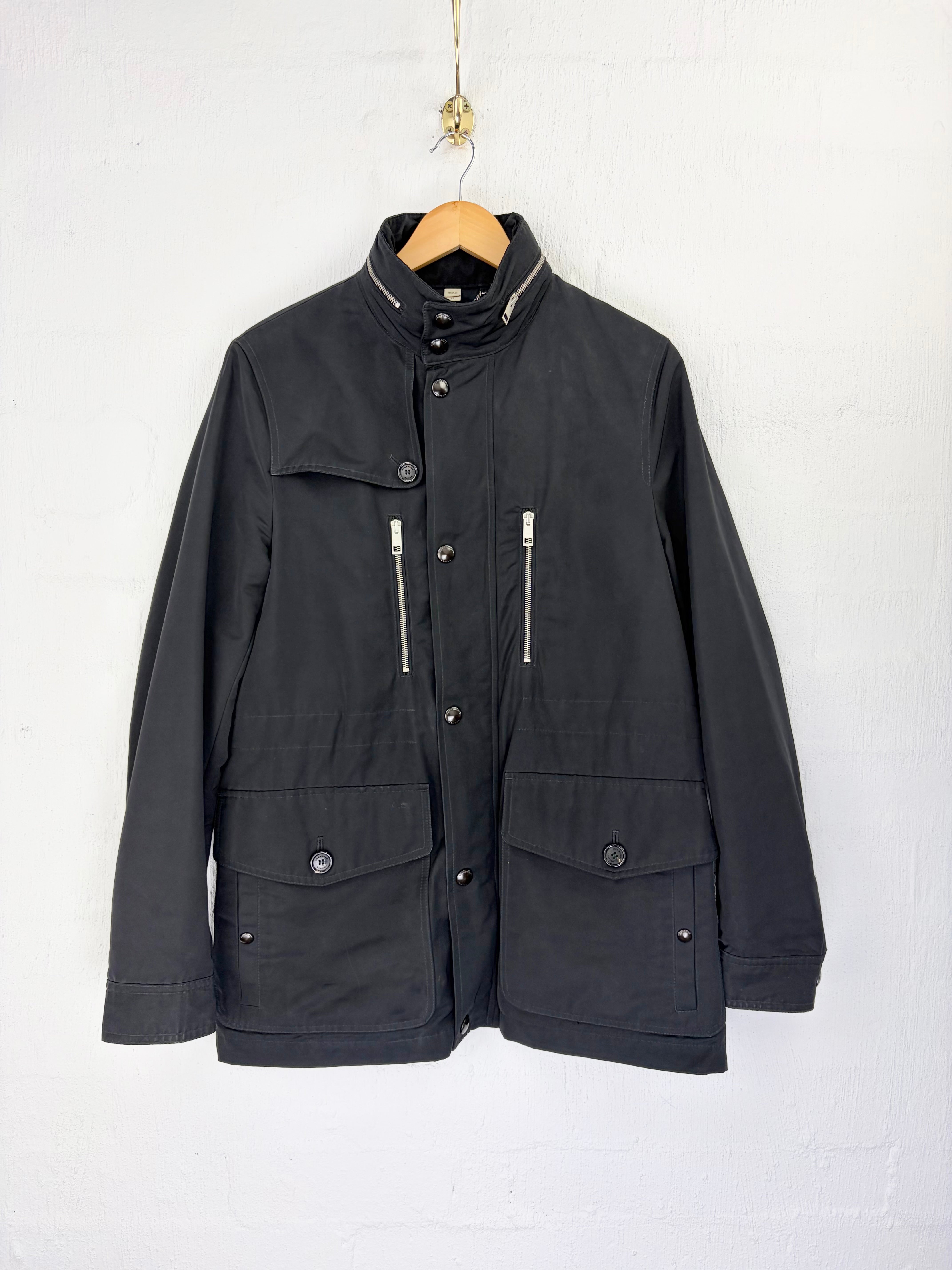 A black utility jacket with silver zipper details, hanging on a wooden hanger against a white wall.