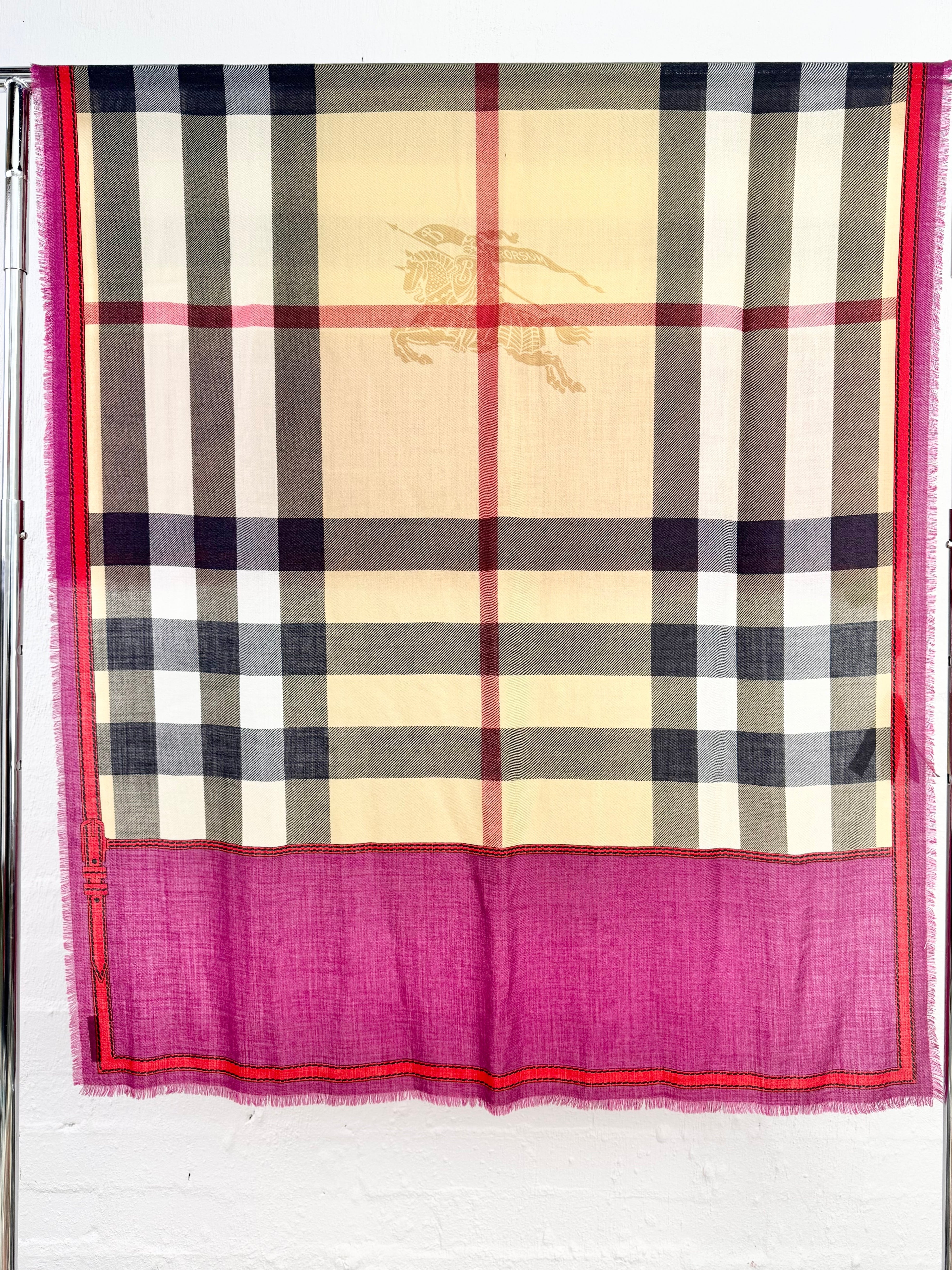 A Burberry check scarf with a vibrant magenta pink border, hanging on a metal rack against a white brick wall. The scarf features the classic beige, black, and white check pattern with a gold equestrian logo, and a solid magenta pink border with red and gold stripes.