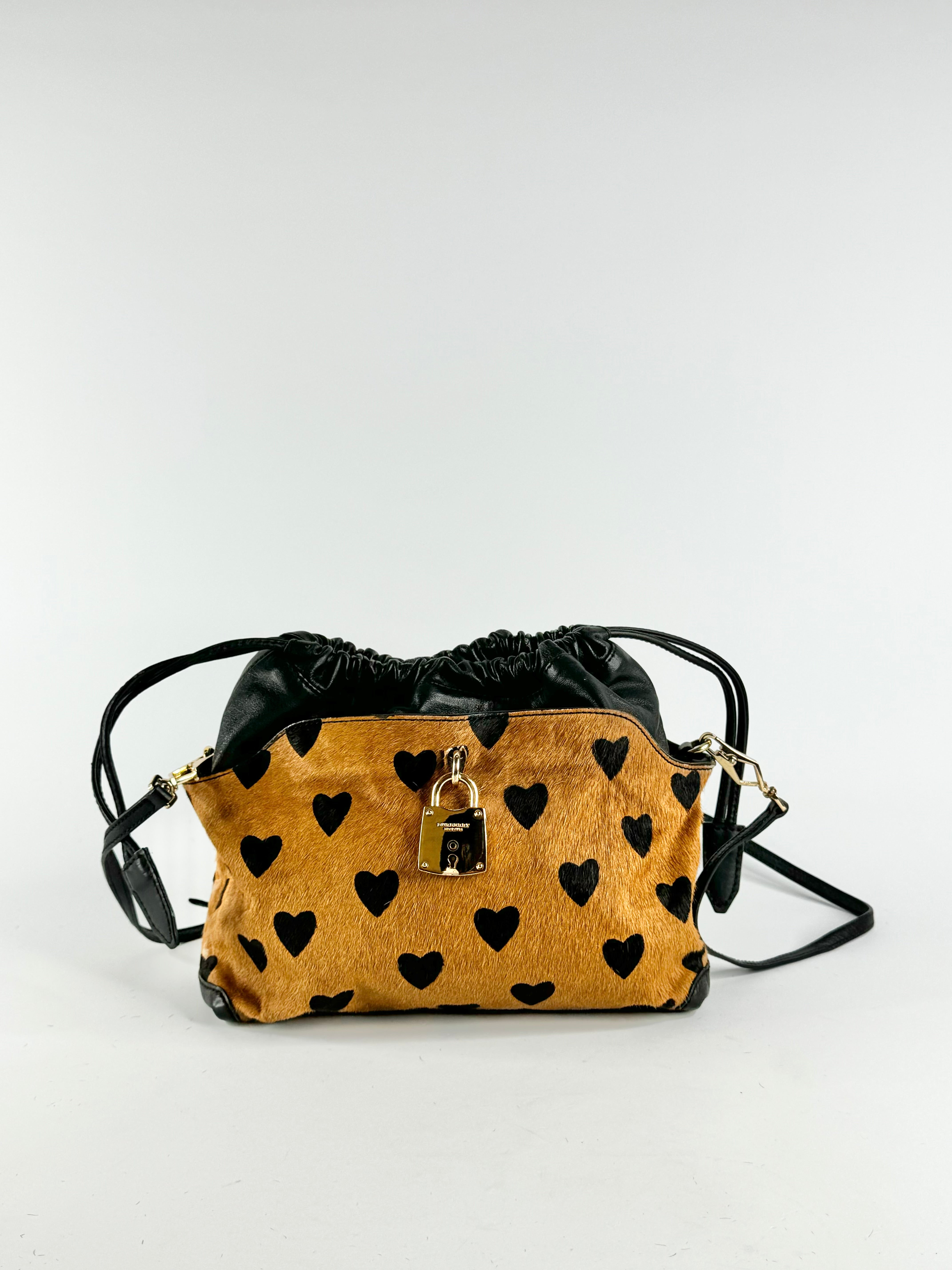 A small crossbody bag with a mustard yellow and black heart-patterned front, black leather top and trim, and a gold padlock detail, shown on a plain light gray background.
