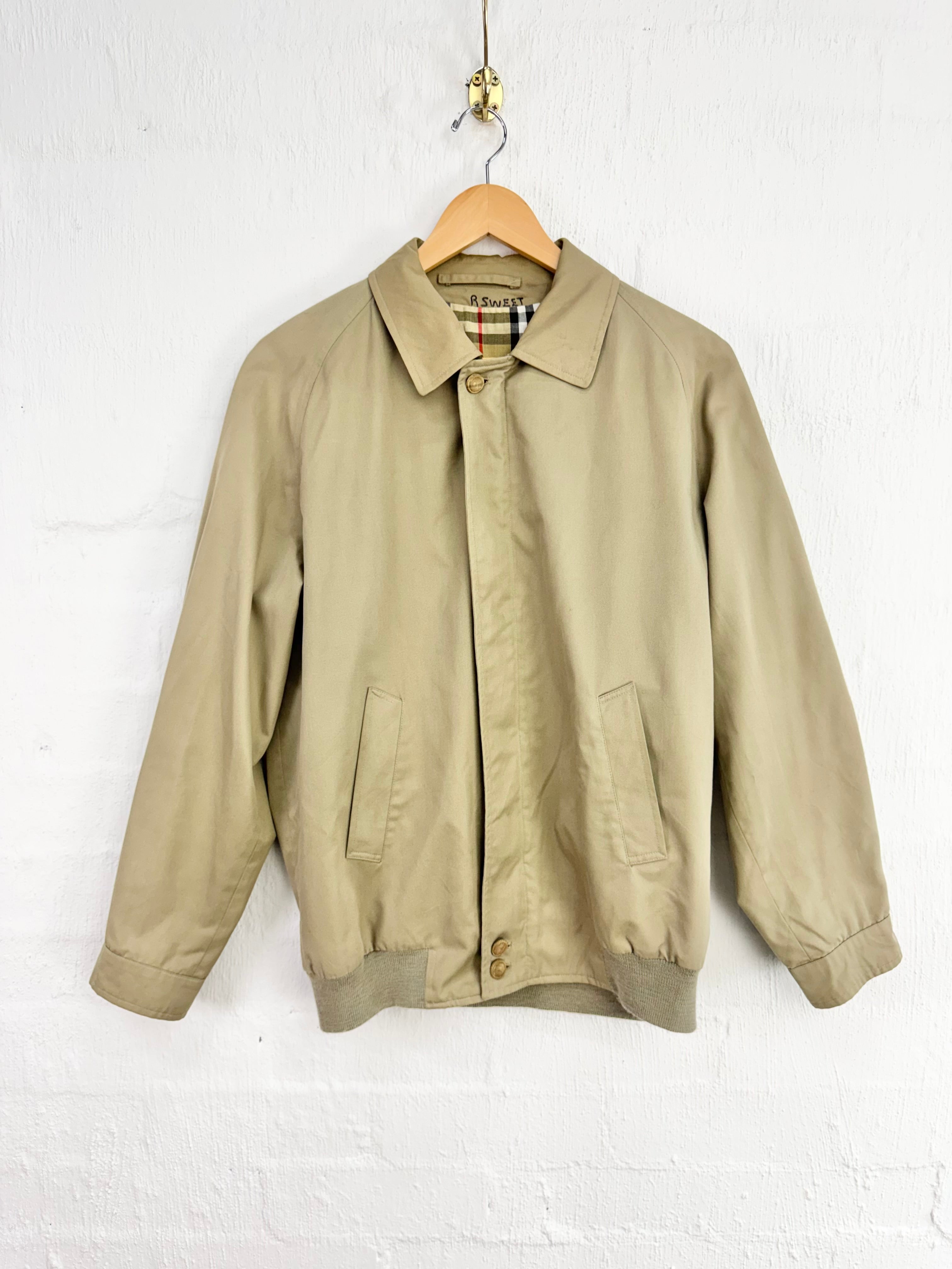 A beige Burberry jacket hanging on a wooden hanger against a white painted brick wall.