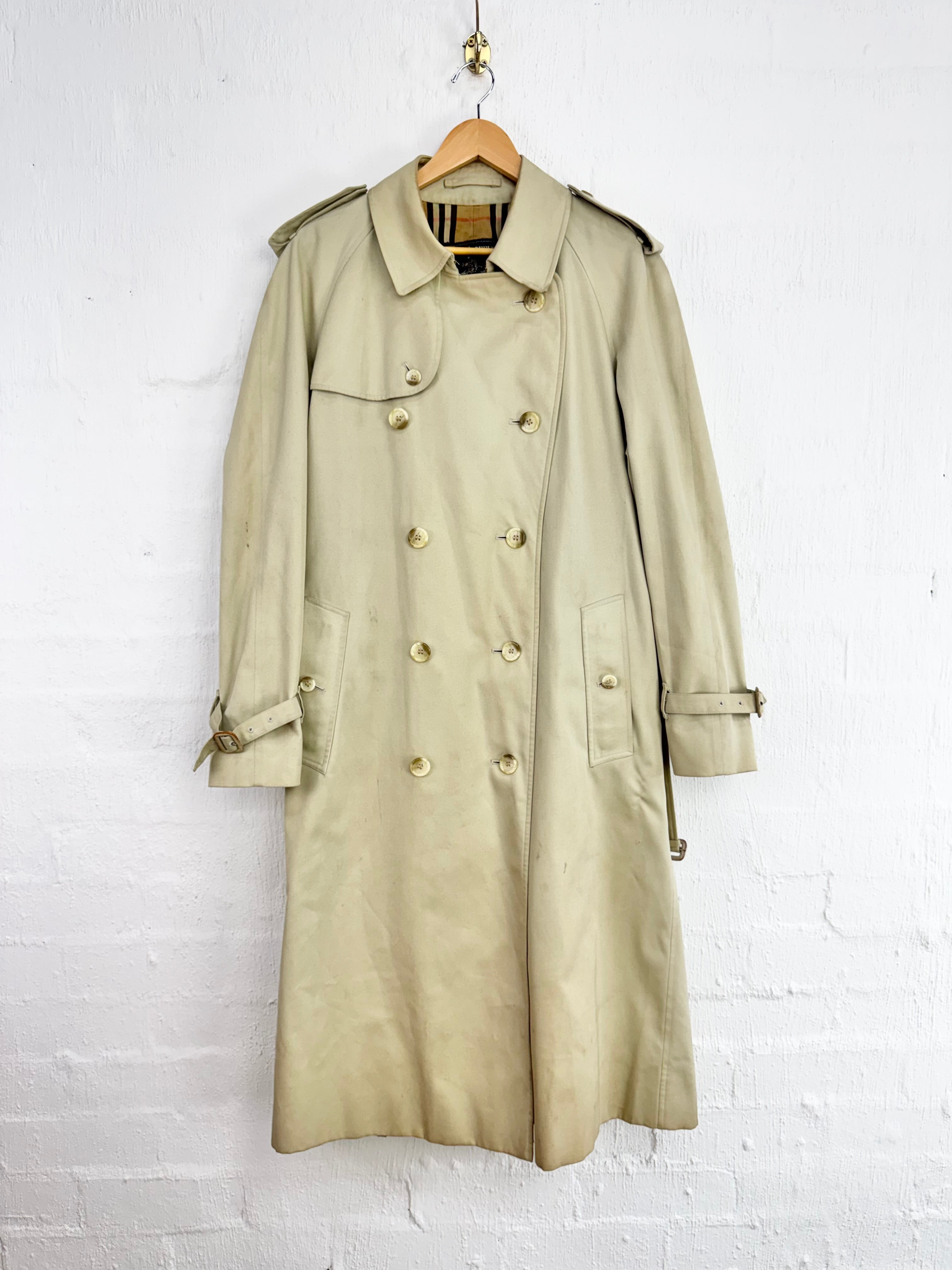 A classic beige trench coat hanging on a wooden hanger against a white painted brick wall.