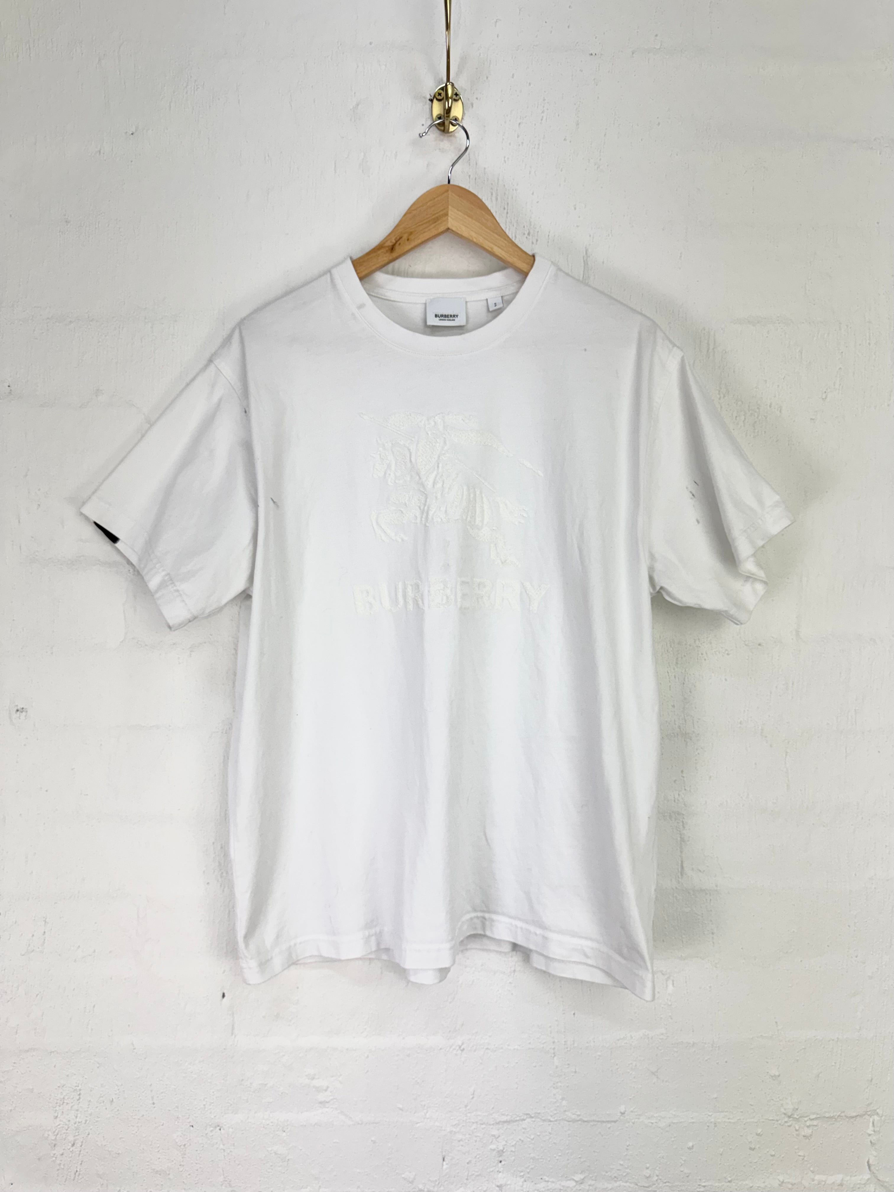 A white short-sleeve crewneck t-shirt with a tonal, slightly faded graphic on the front, hanging on a wooden hanger against a white painted brick wall.
