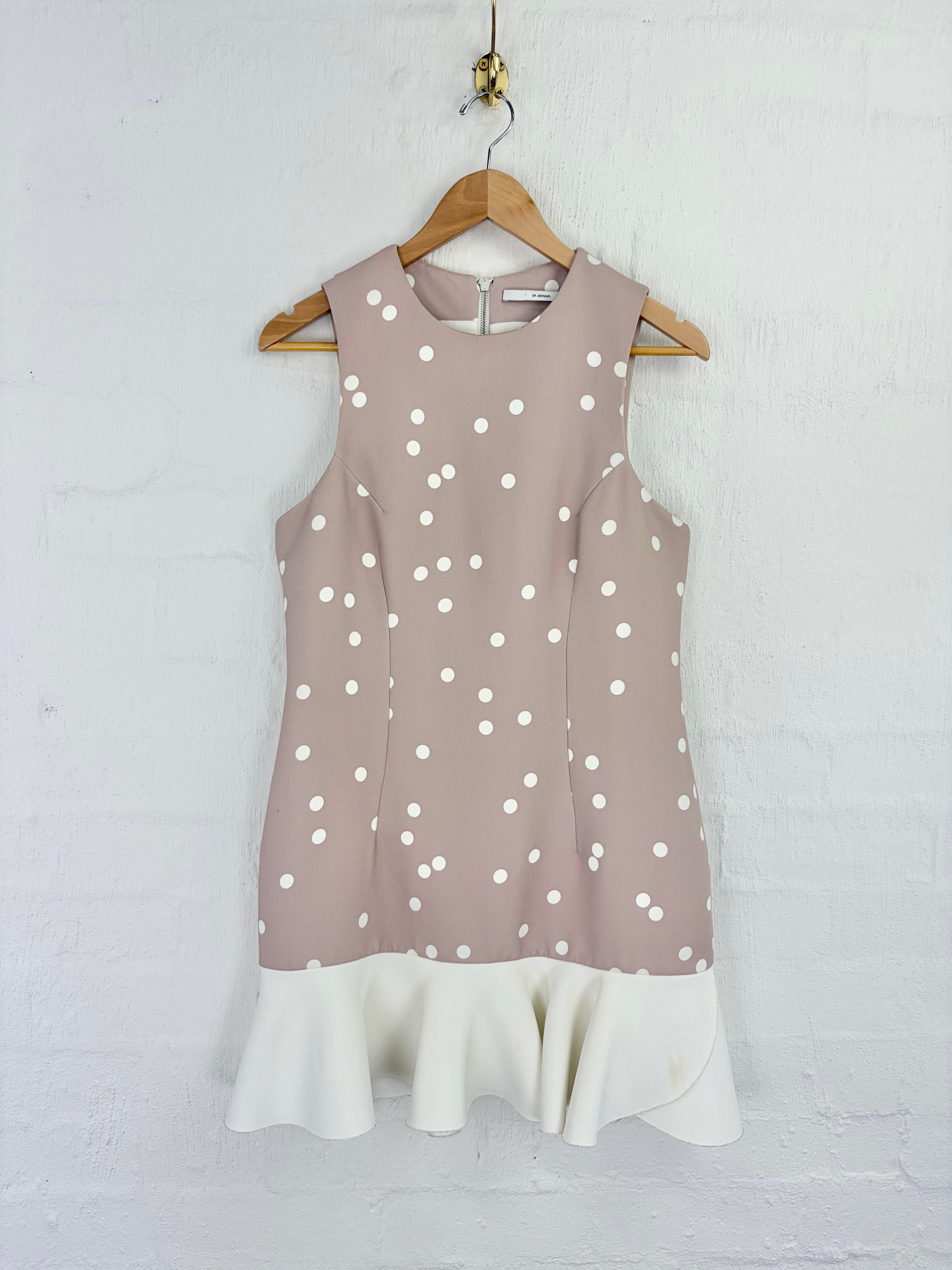A sleeveless dusty pink dress with white polka dots and a contrasting white ruffled hem, hanging on a wooden hanger against a white painted brick wall.