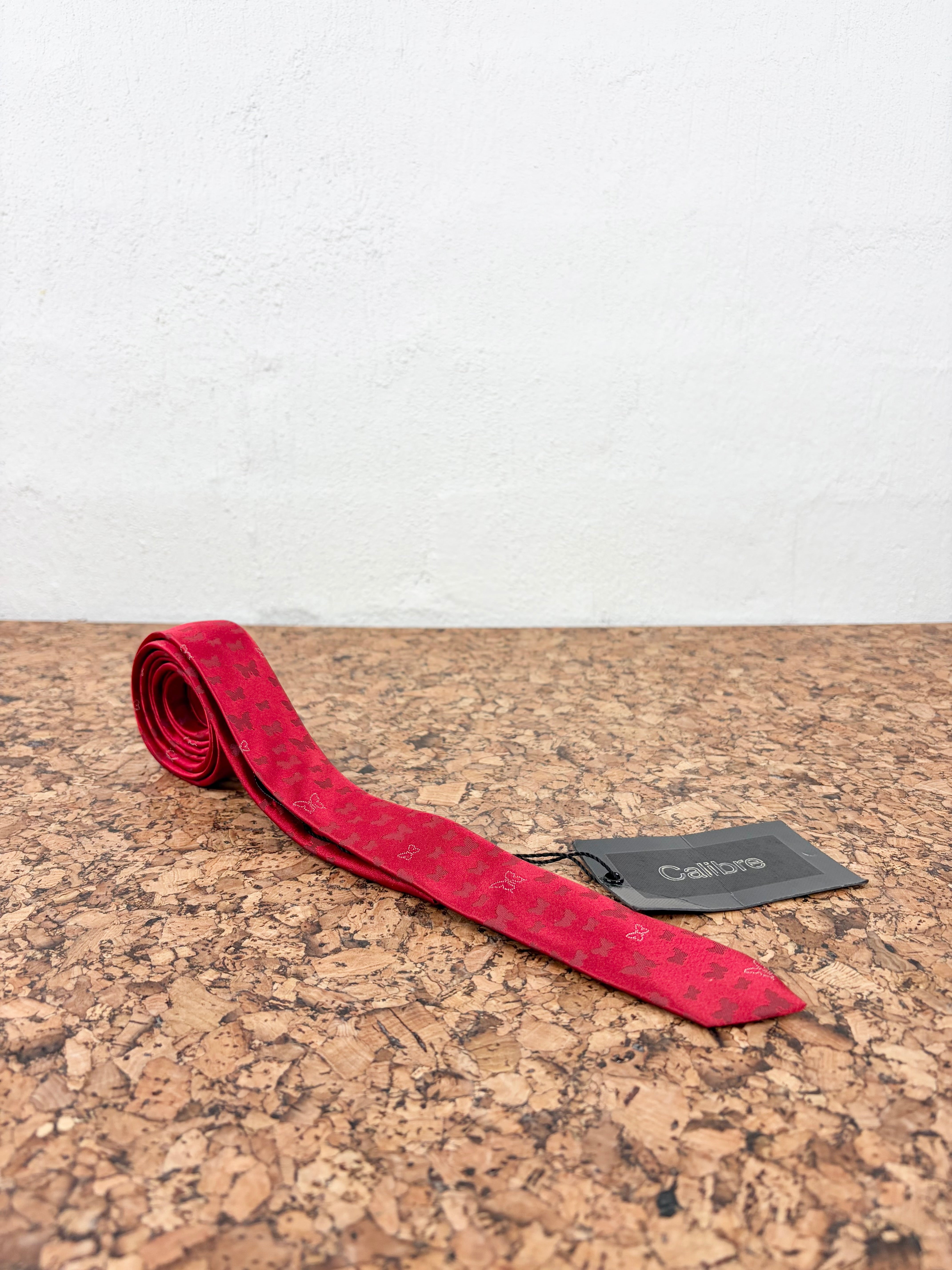 A red silk necktie with a subtle repeating geometric pattern, laid out on a brown cork surface against a white textured wall. A black brand tag for 'Calibres' is attached to the tie.