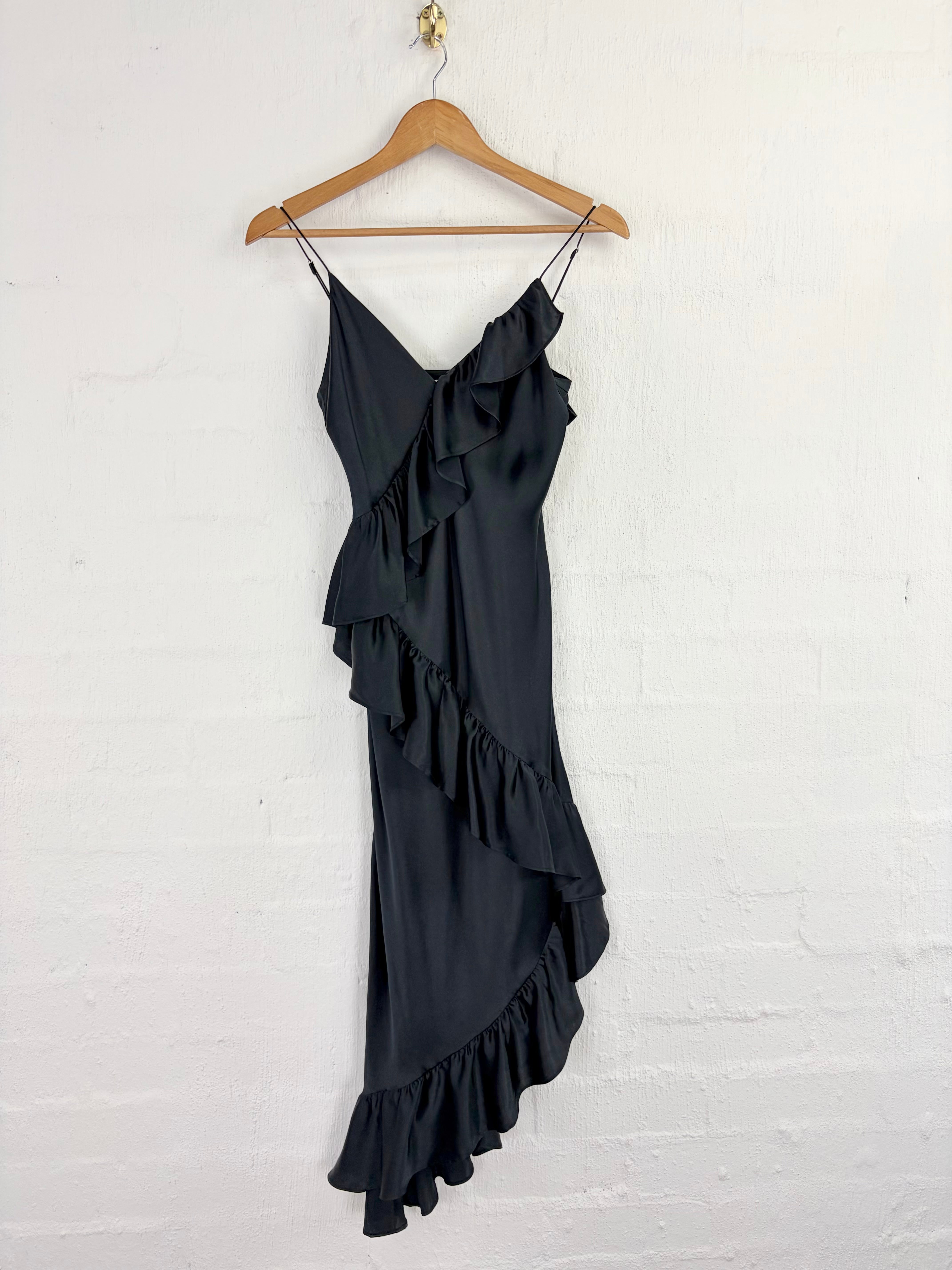 A black ruffled slip dress with thin spaghetti straps, hanging on a wooden hanger against a white painted brick wall.