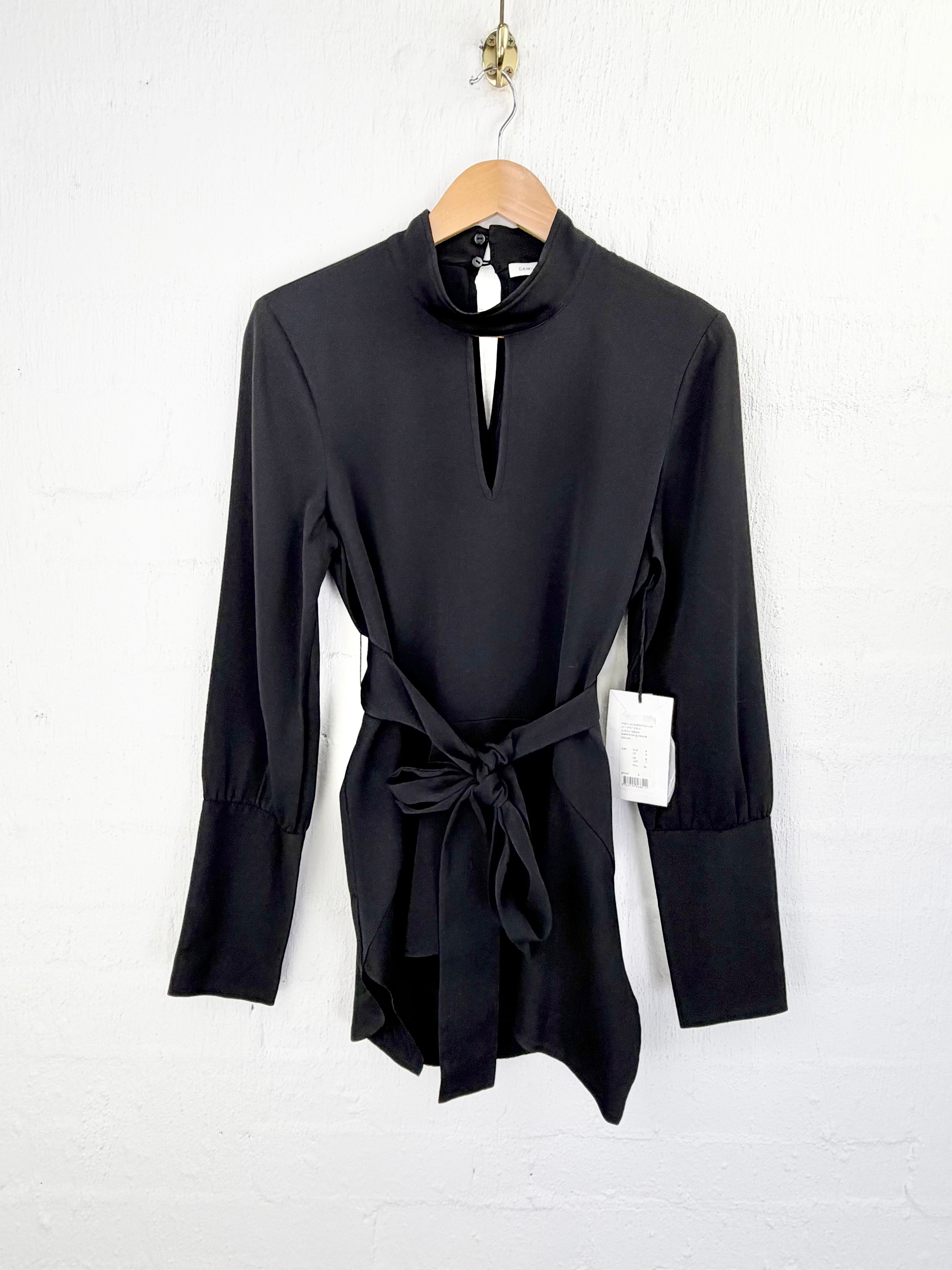 A black long-sleeve wrap-style blouse with a mock neck and self-tie waist belt, hanging on a wooden hanger against a white painted brick wall.