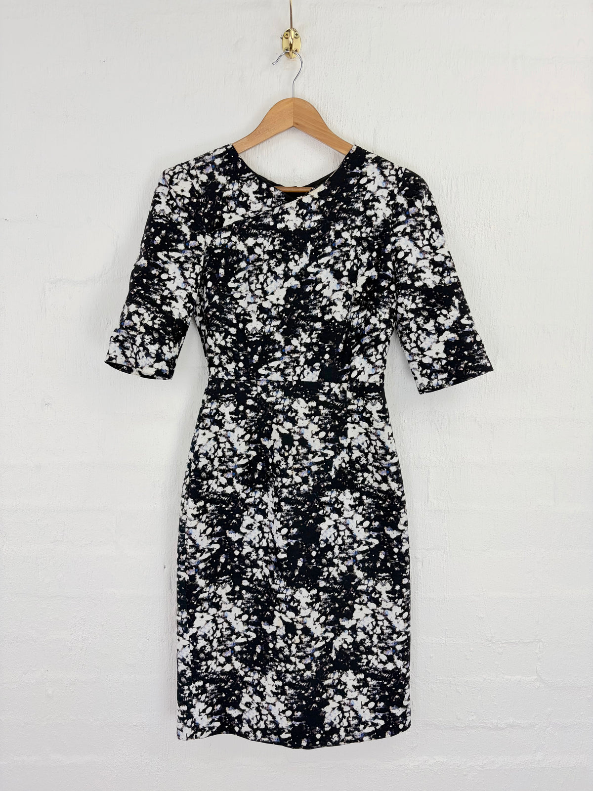 A black and white speckled patterned dress with elbow-length sleeves, hanging on a wooden hanger against a white painted brick wall.