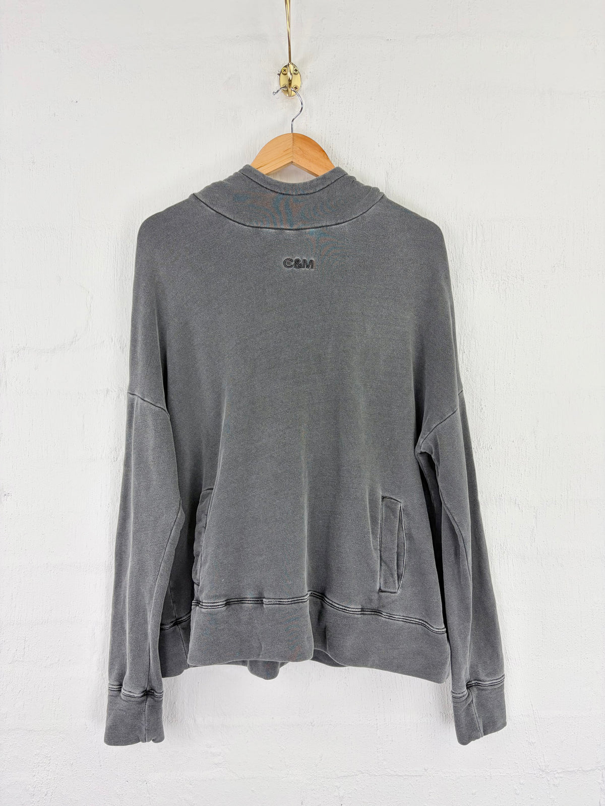 A gray washed long-sleeve sweatshirt with a mock neck, hanging on a wooden hanger against a white brick wall. The brand name 'C.