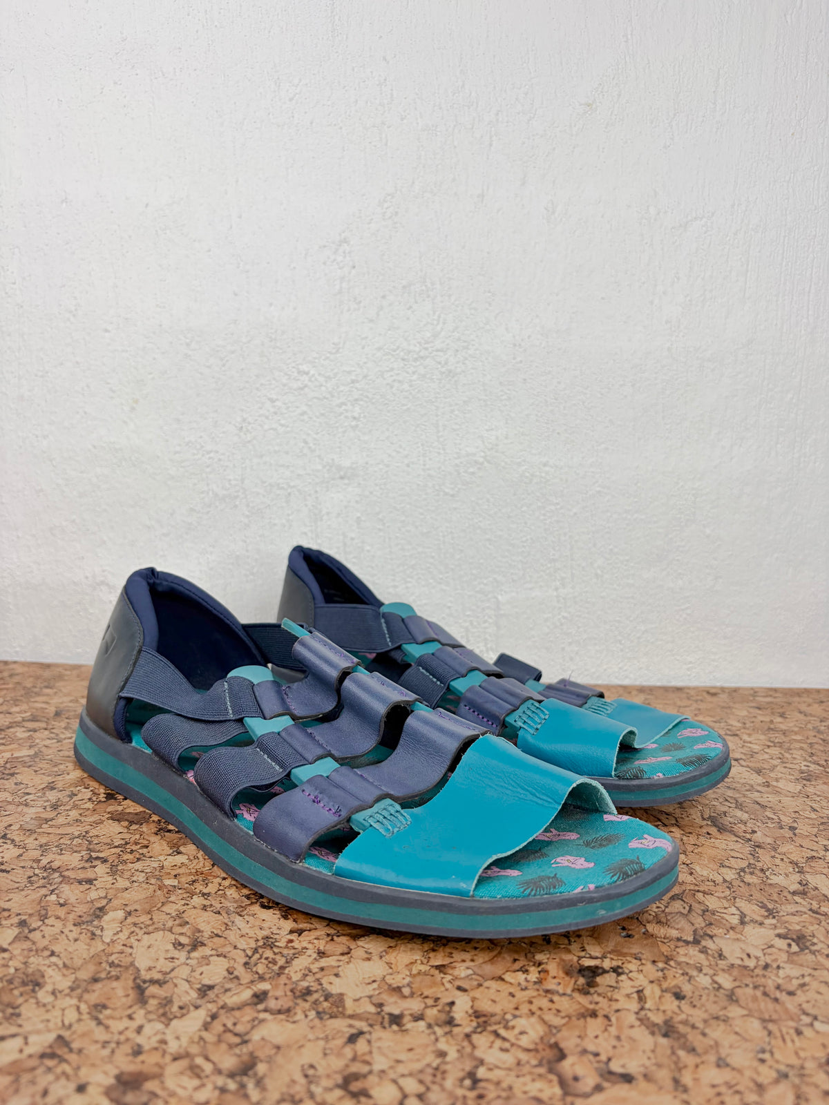 A pair of outdoor slide sandals with teal and navy blue straps and soles, resting on a textured cork surface against a white wall.