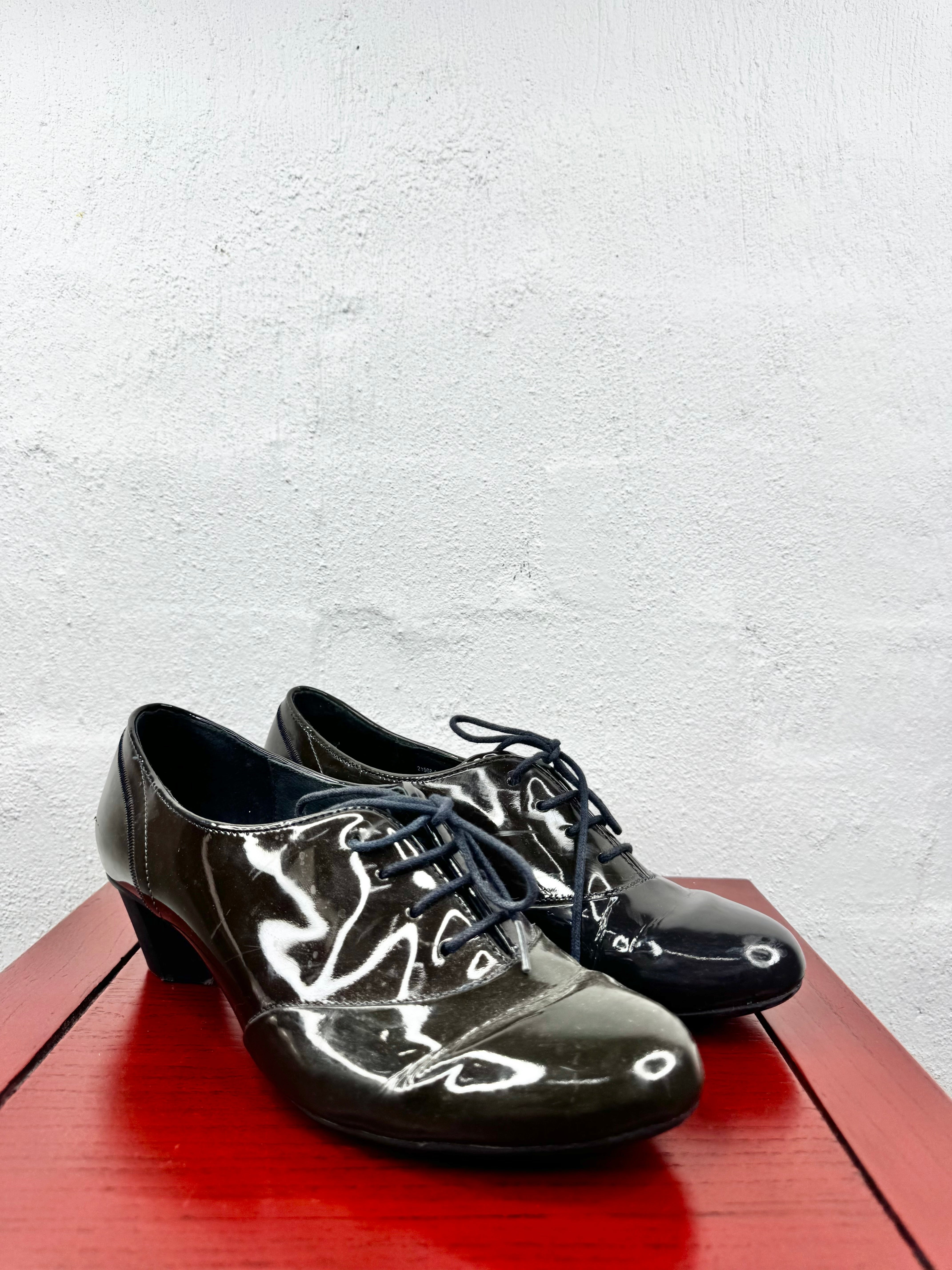 A pair of glossy black lace-up dance shoes with low block heels, displayed on a red wooden surface against a textured white wall.