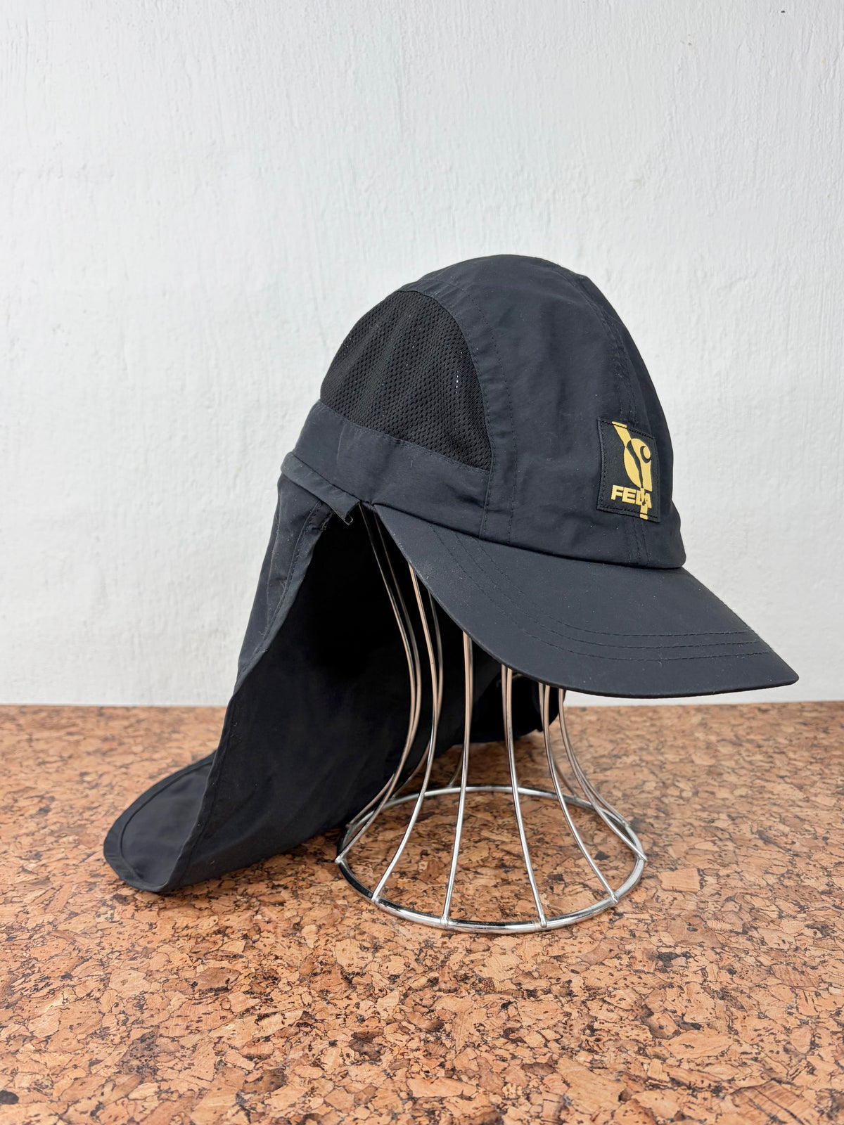 A black sun hat with a neck flap and side mesh ventilation, featuring a small yellow logo on the side, displayed on a wire stand against a white wall.