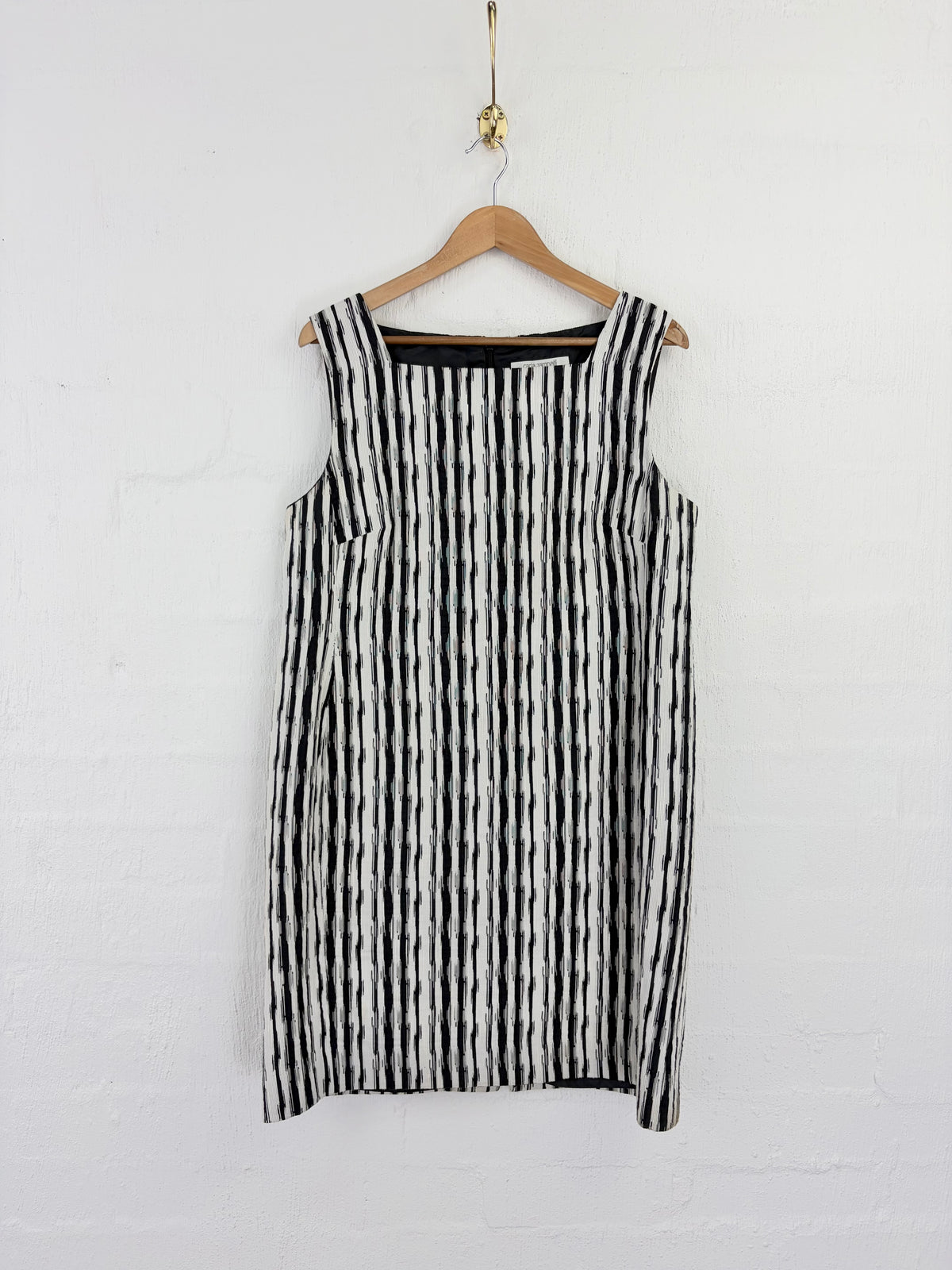 A sleeveless black and white vertically striped dress hanging on a wooden hanger against a white painted brick wall.