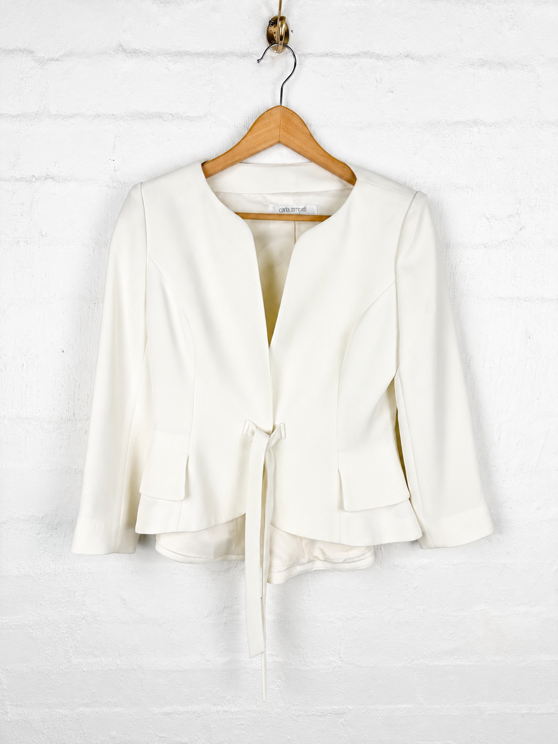 An off-white wrap jacket with three-quarter sleeves, displayed on a wooden hanger against a white brick wall.