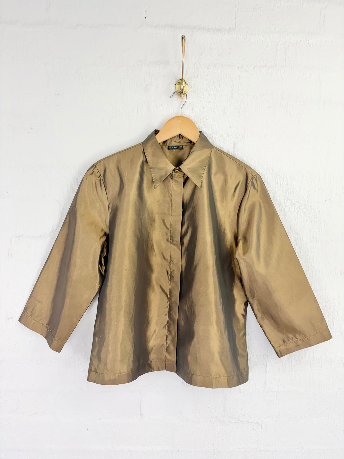A metallic gold long-sleeve button-up shirt hanging on a wooden hanger against a white painted brick wall.