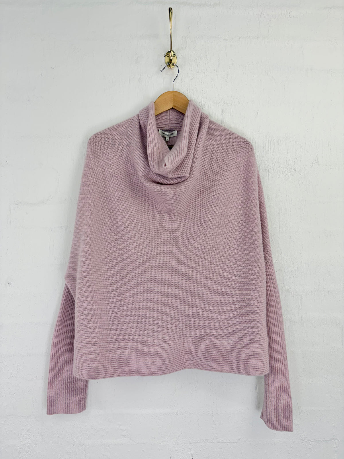 A dusty pink ribbed knit cowl neck sweater hanging on a wooden hanger against a white painted brick wall.
