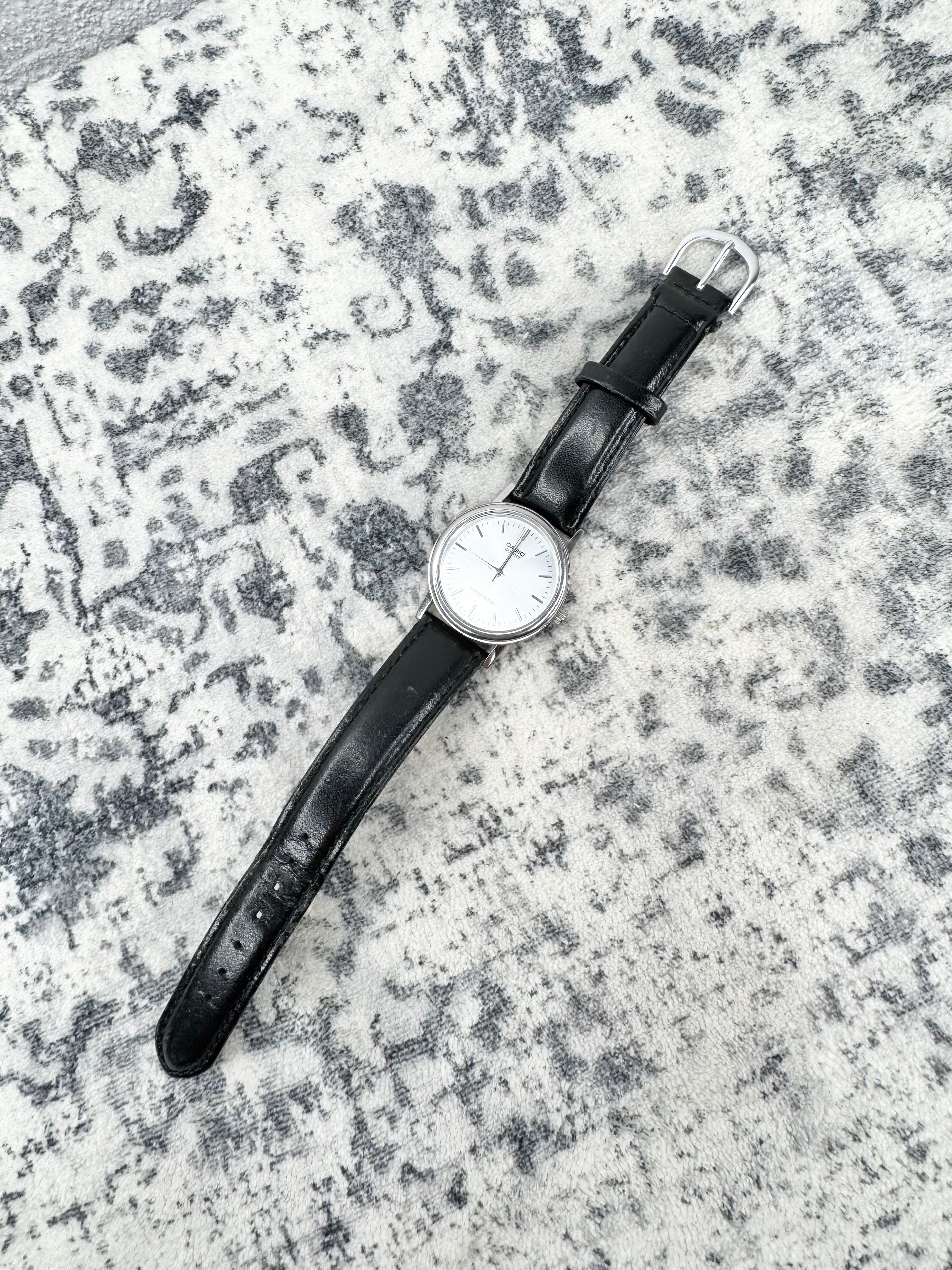 A classic wristwatch with a black leather strap and a silver-toned round dial, laid diagonally on a textured gray and white patterned rug.