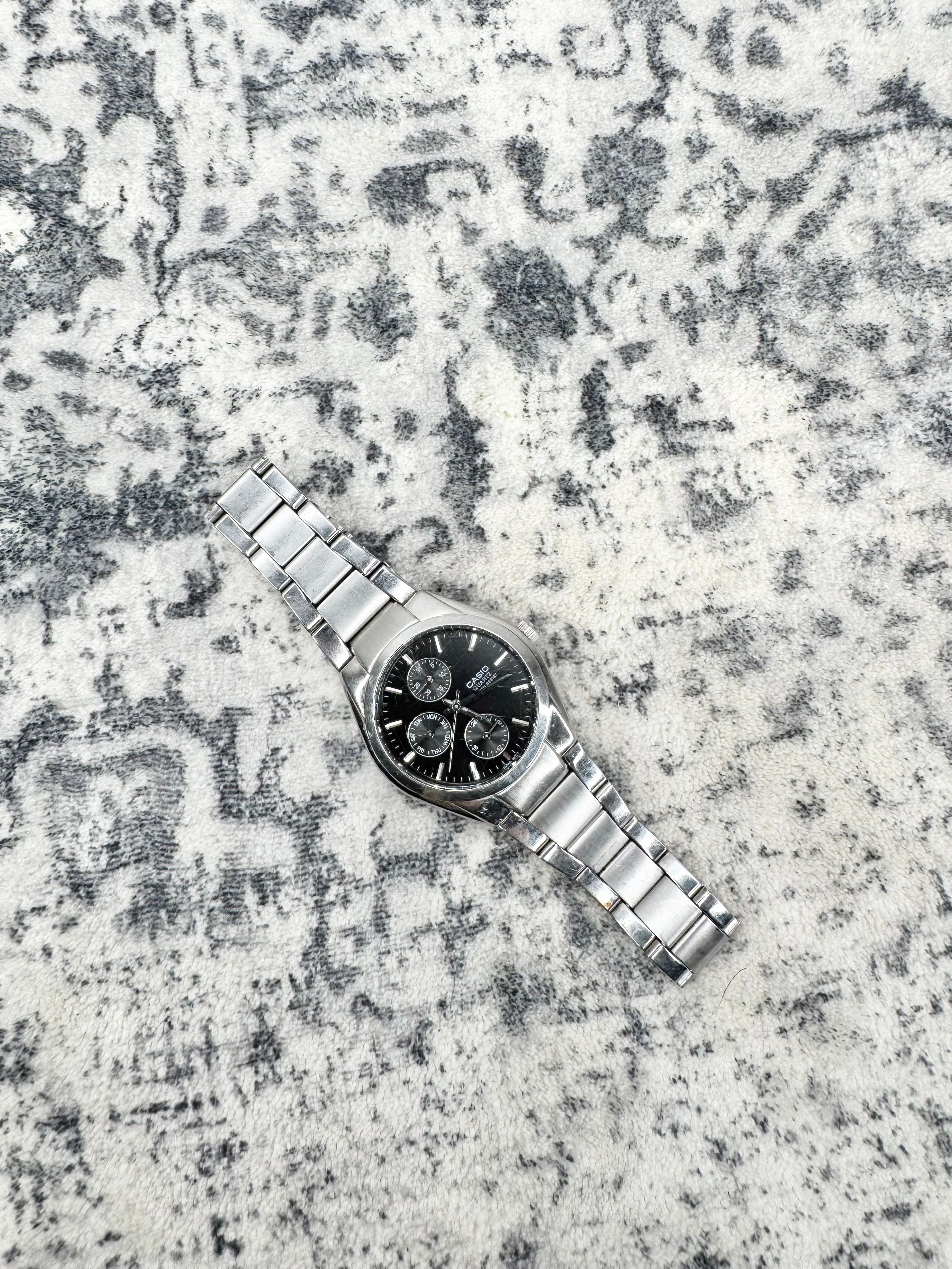 A silver stainless steel wristwatch with a black dial and three subdials, laid diagonally on a textured gray and white patterned surface.