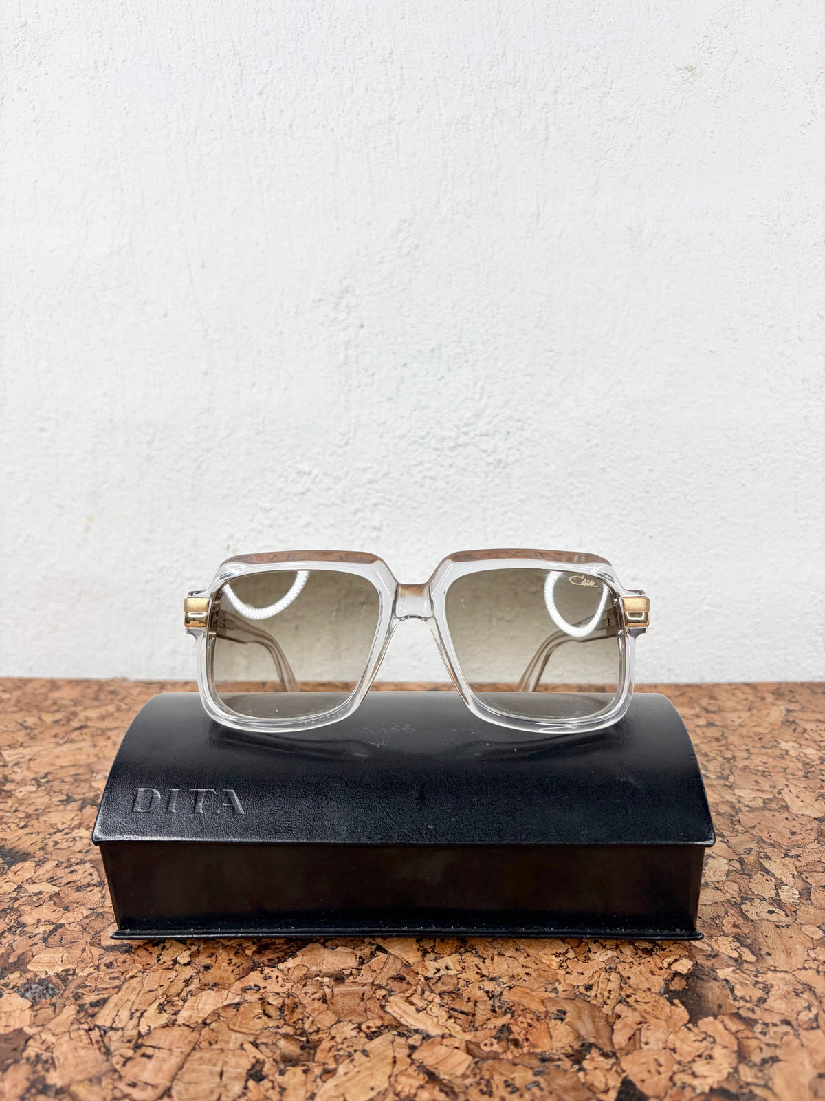 A pair of DITA brand square sunglasses with clear frames and gold-tone hinges, resting on a black DITA branded case on a cork surface against a white wall.