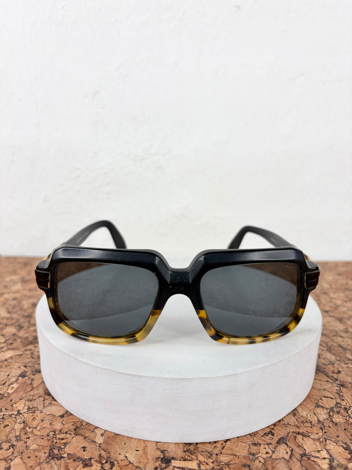 Square sunglasses with thick black frames and dark tortoiseshell-patterned rims, resting on a white cylindrical pedestal over a cork surface against a white wall.