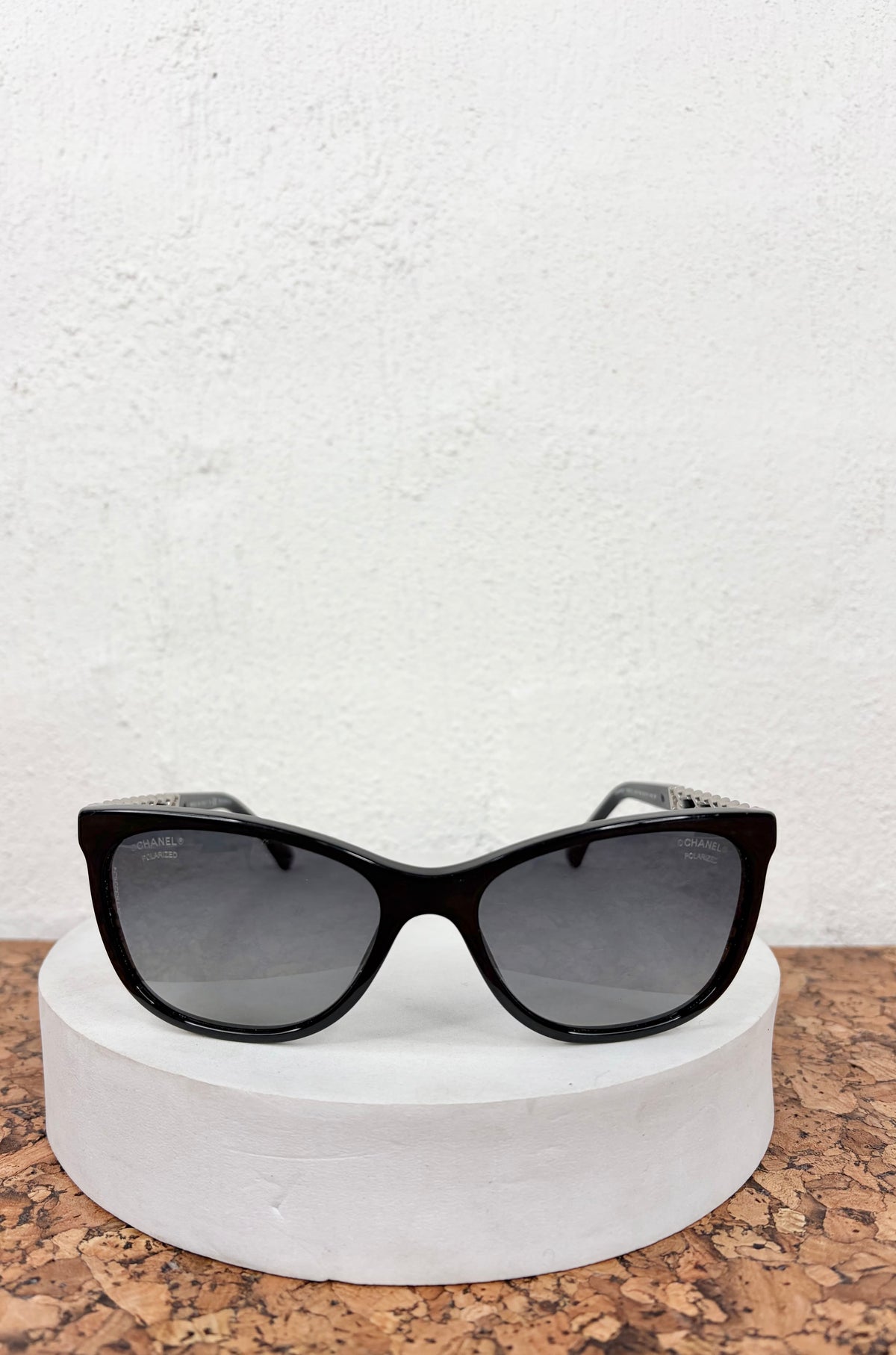 Black cat-eye sunglasses with dark gray lenses, displayed on a round white pedestal against a textured white wall and cork surface.