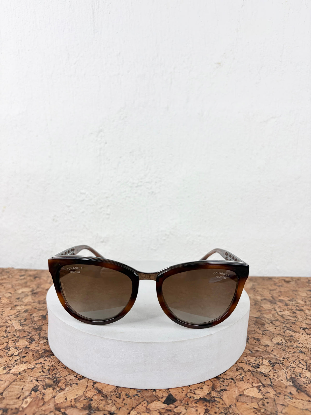 A pair of cat-eye sunglasses with dark brown tortoiseshell frames and brown-tinted lenses, displayed on a round white pedestal against a white wall.