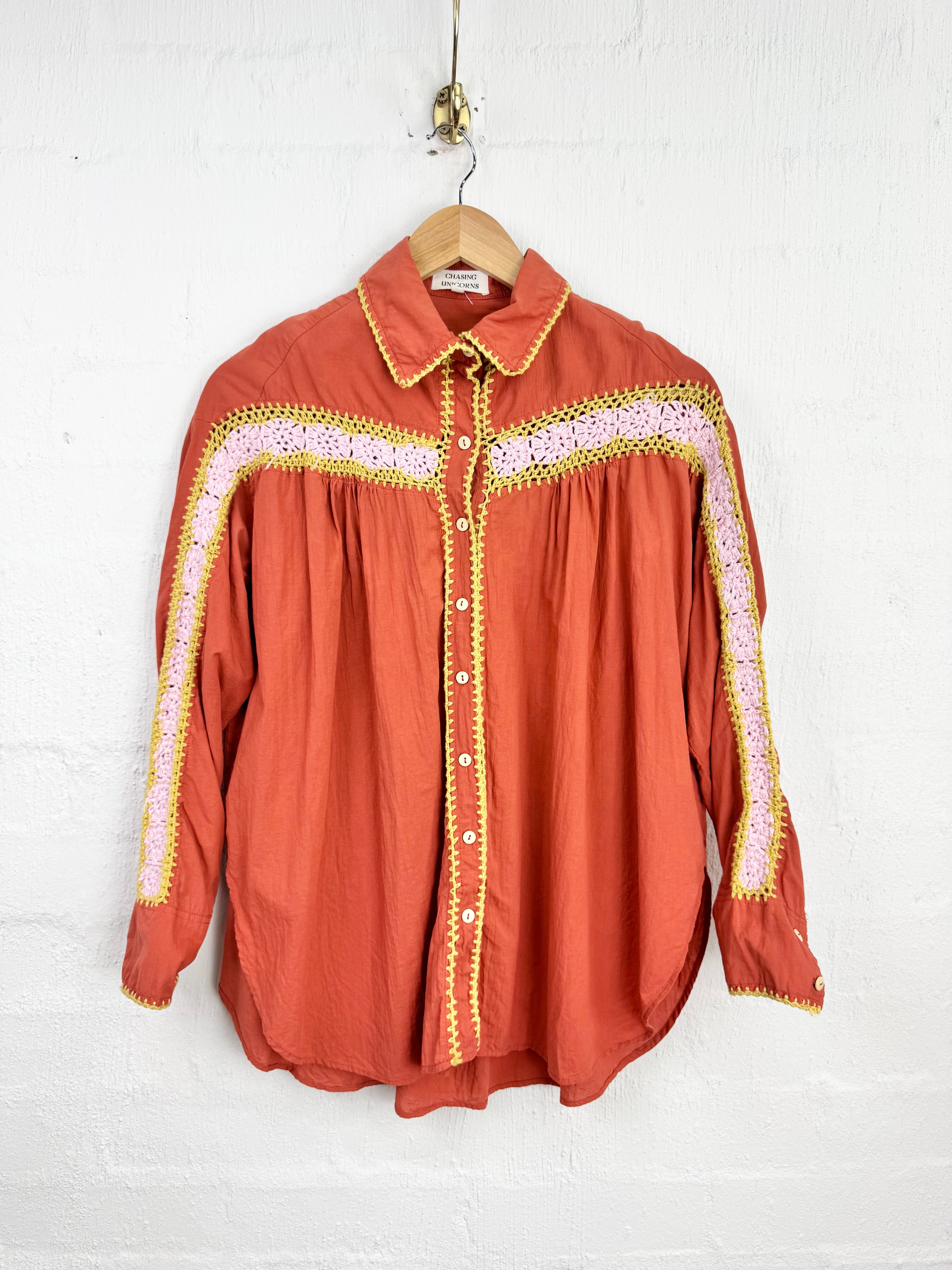 A rust-orange western-style blouse with yellow and pink embroidered details, hanging on a wooden hanger against a white brick wall.