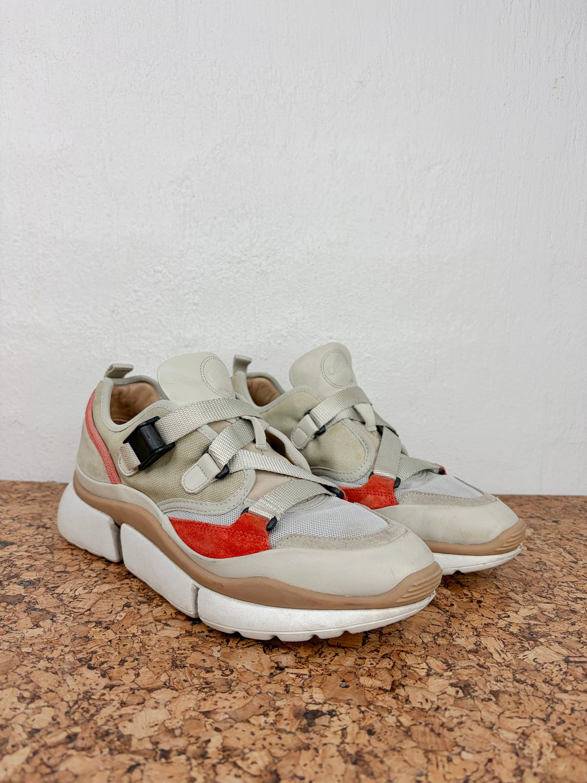 A pair of chunky low-top sneakers in light beige and gray suede and mesh, featuring bright red side panels and brown midsoles, displayed on a cork surface against a white wall.