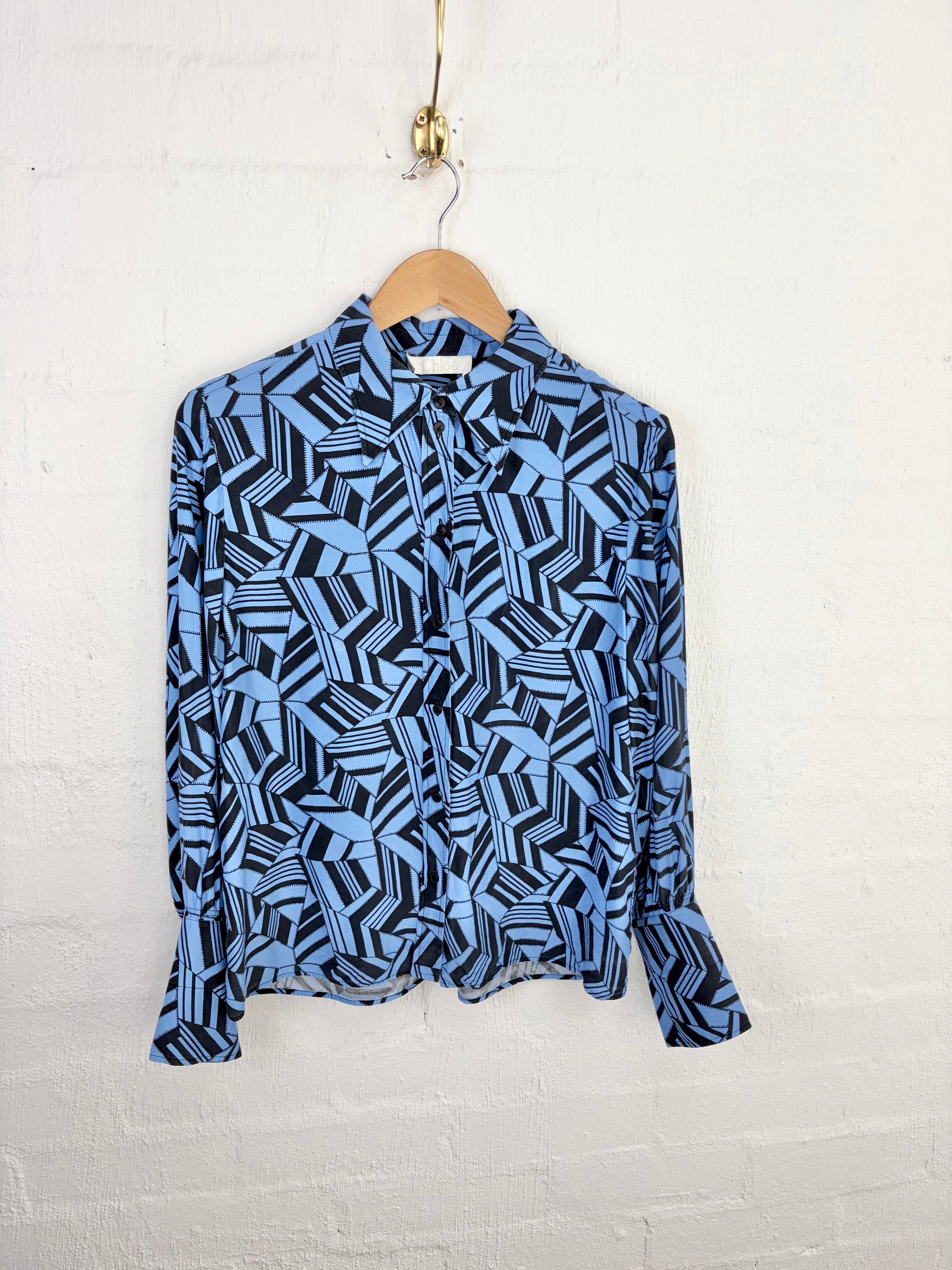 A long-sleeve button-up blouse with an abstract geometric pattern in light blue, navy, and black, hanging on a wooden hanger against a white painted brick wall.