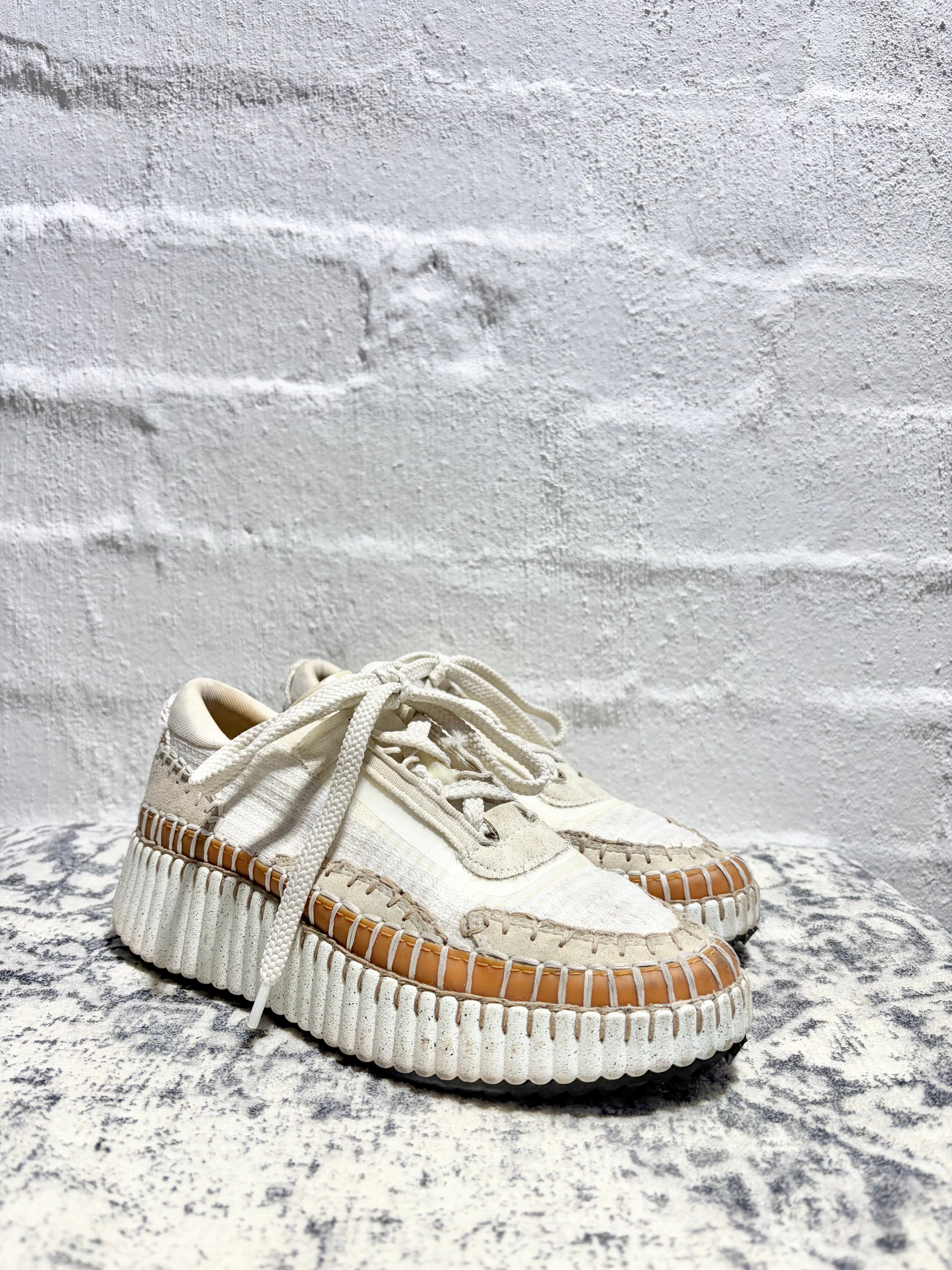 A pair of chunky low-top platform sneakers in off-white and beige, featuring textured woven panels, thick ridged white platform soles, and tan leather trim, displayed against a white brick wall.