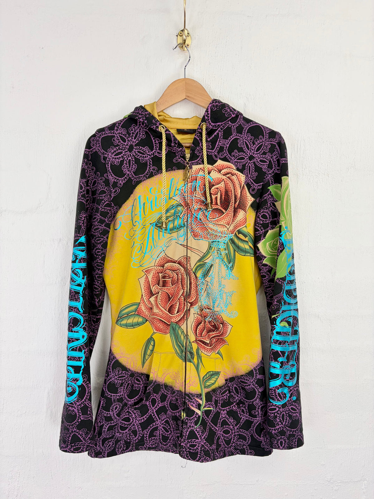 A vintage Grungecore-style zip-up hoodie hanging on a wooden hanger against a white brick wall. The hoodie features a dark purple lace-like pattern on the sleeves and hem, a large yellow circular graphic on the front with three pinkish-brown roses and turquoise text, and blue 'Grungecore' text printed down the sleeves.