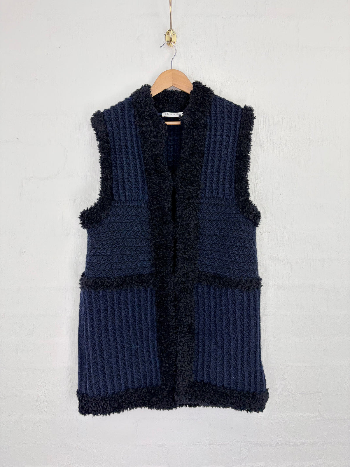 A sleeveless navy blue knit vest with a dark fuzzy trim, hanging on a wooden hanger against a white painted brick wall.