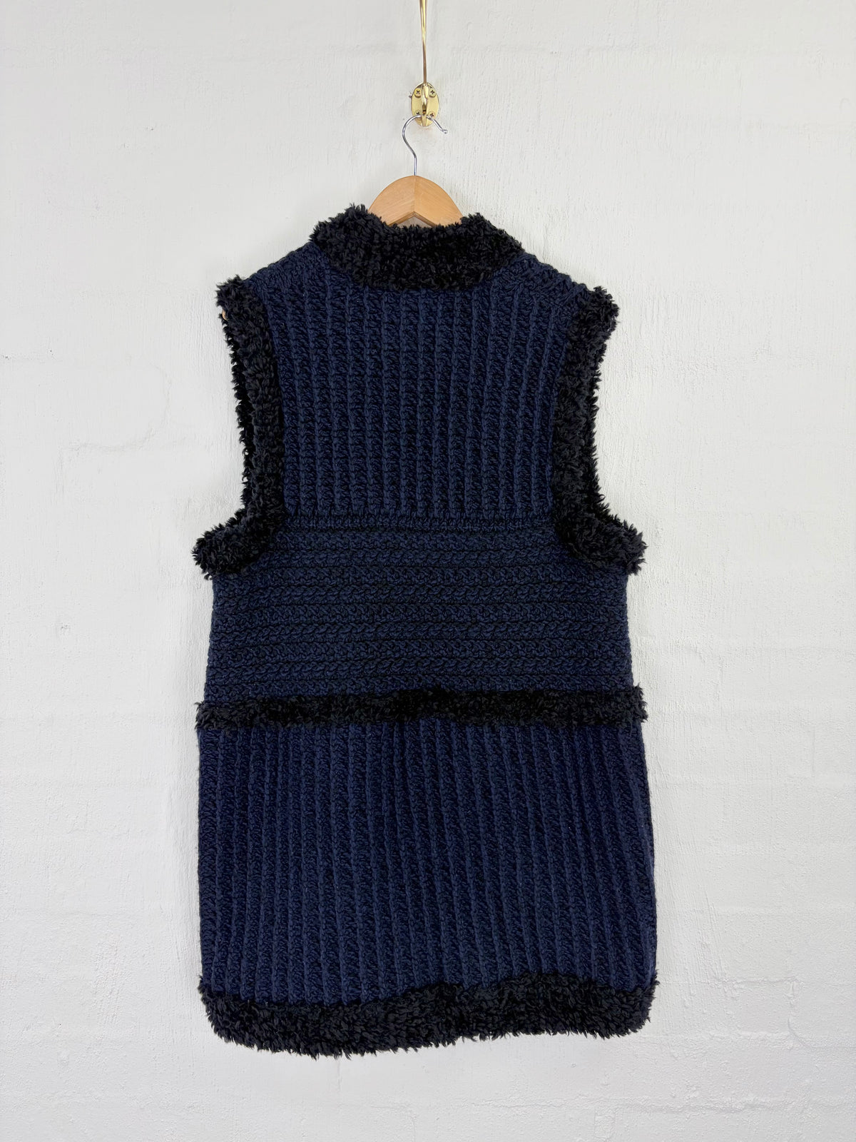 A sleeveless navy blue knit vest with black fuzzy trim, hanging on a wooden hanger against a white painted brick wall.