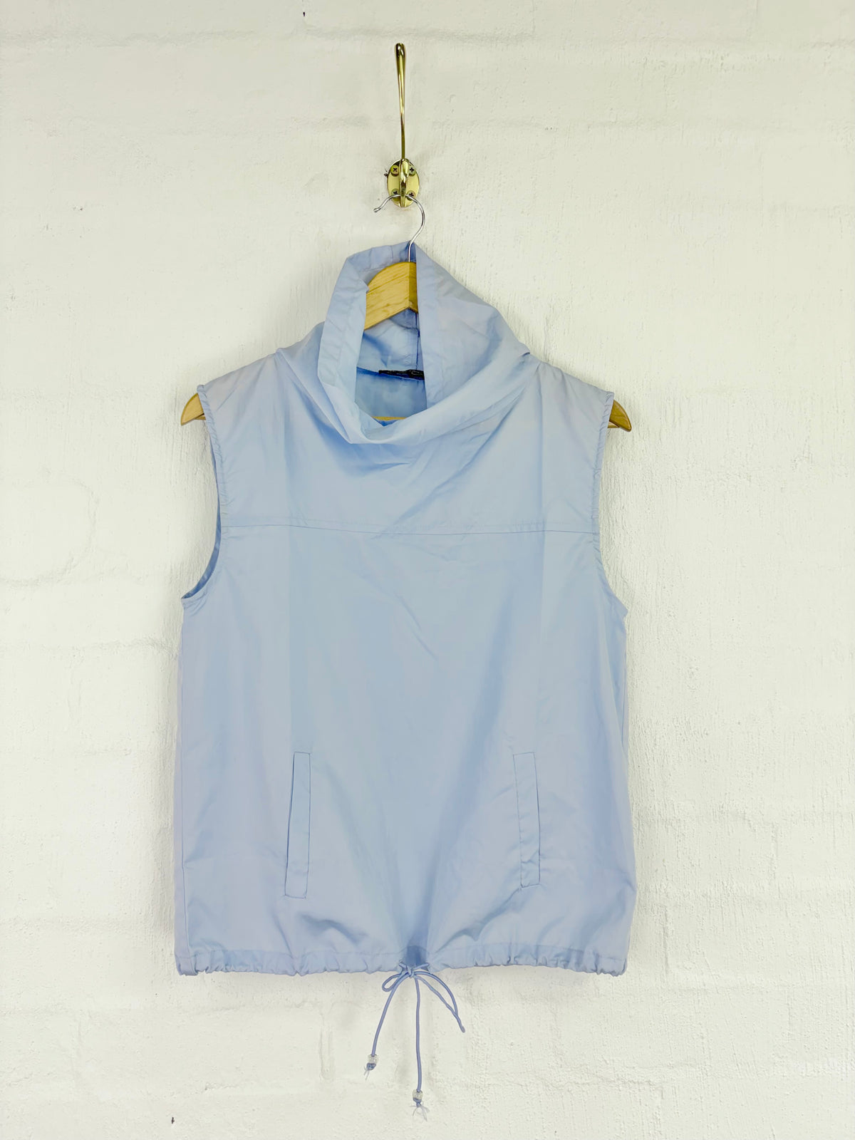 A light blue sleeveless cowl-neck vest with a drawstring hem, hanging on a wooden hanger from a gold hook against a white brick wall.