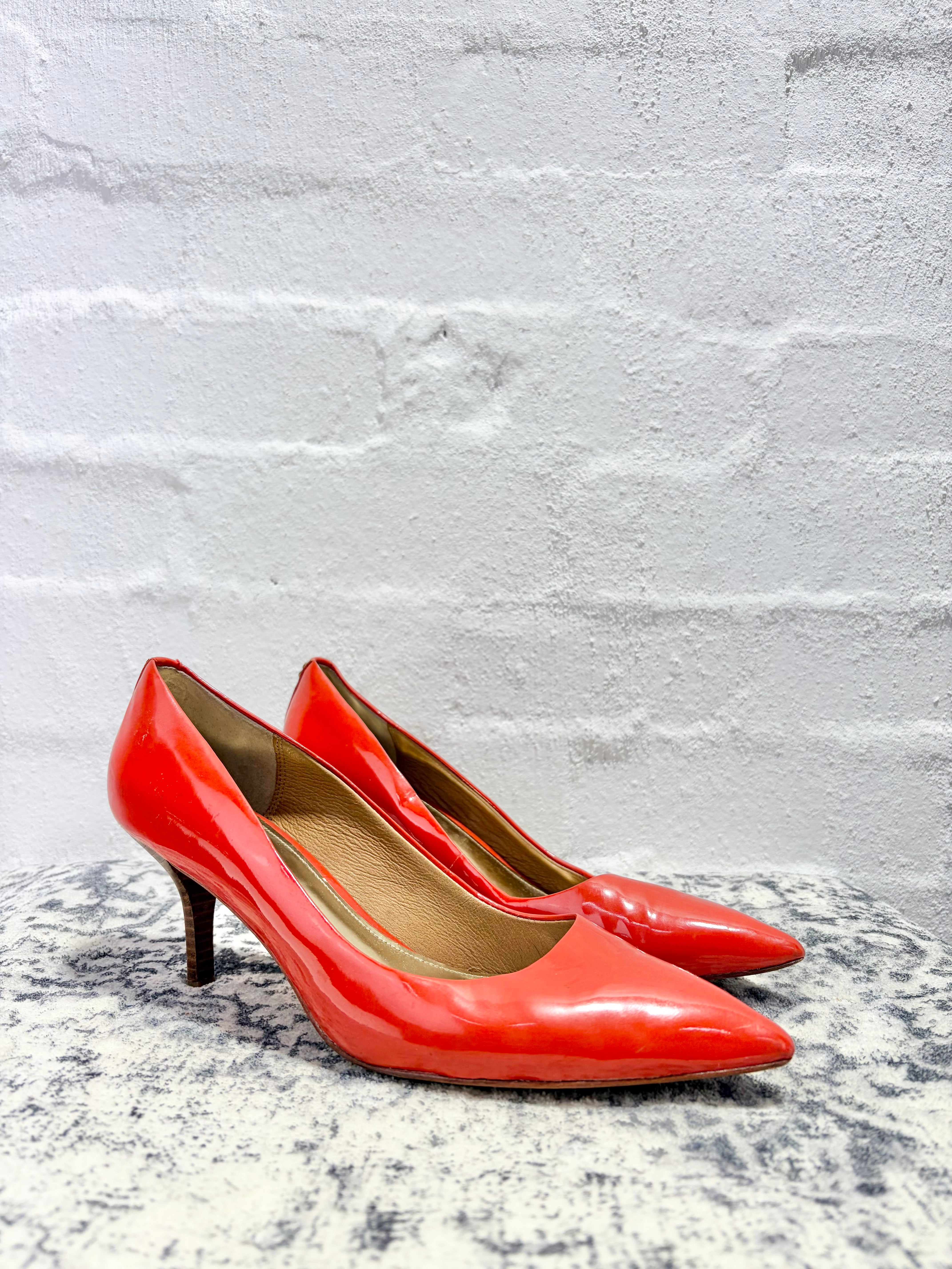 A pair of bright red patent leather kitten heels with pointed toes, displayed on a textured gray surface against a white brick wall background.