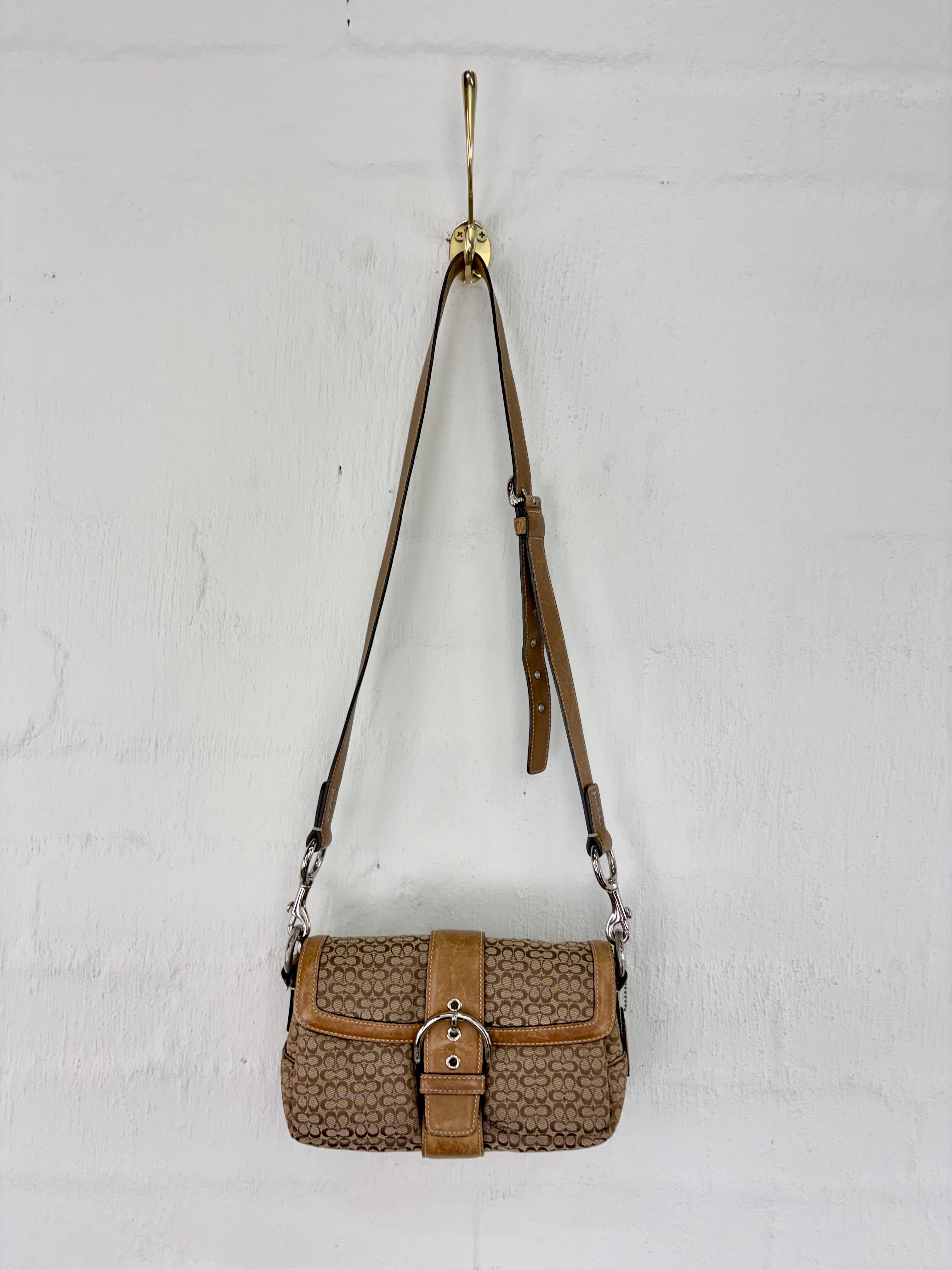 A vintage-style shoulder bag with a brown monogram pattern and tan leather trim, hanging from a gold hook against a white brick wall.