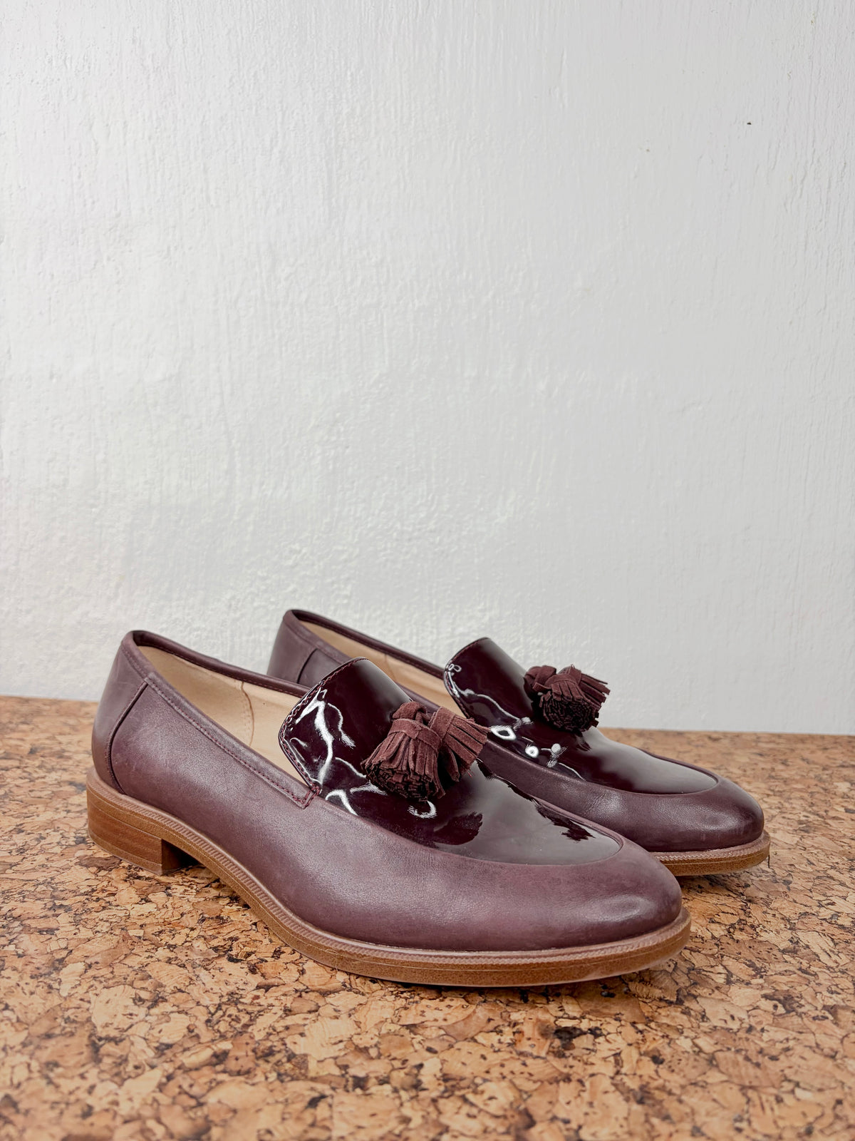 A pair of women's low-heeled loafers in a deep burgundy-brown color, featuring glossy patent leather toe caps, tassel details, and a stacked wood-look heel, displayed on a textured brown surface against a white wall.