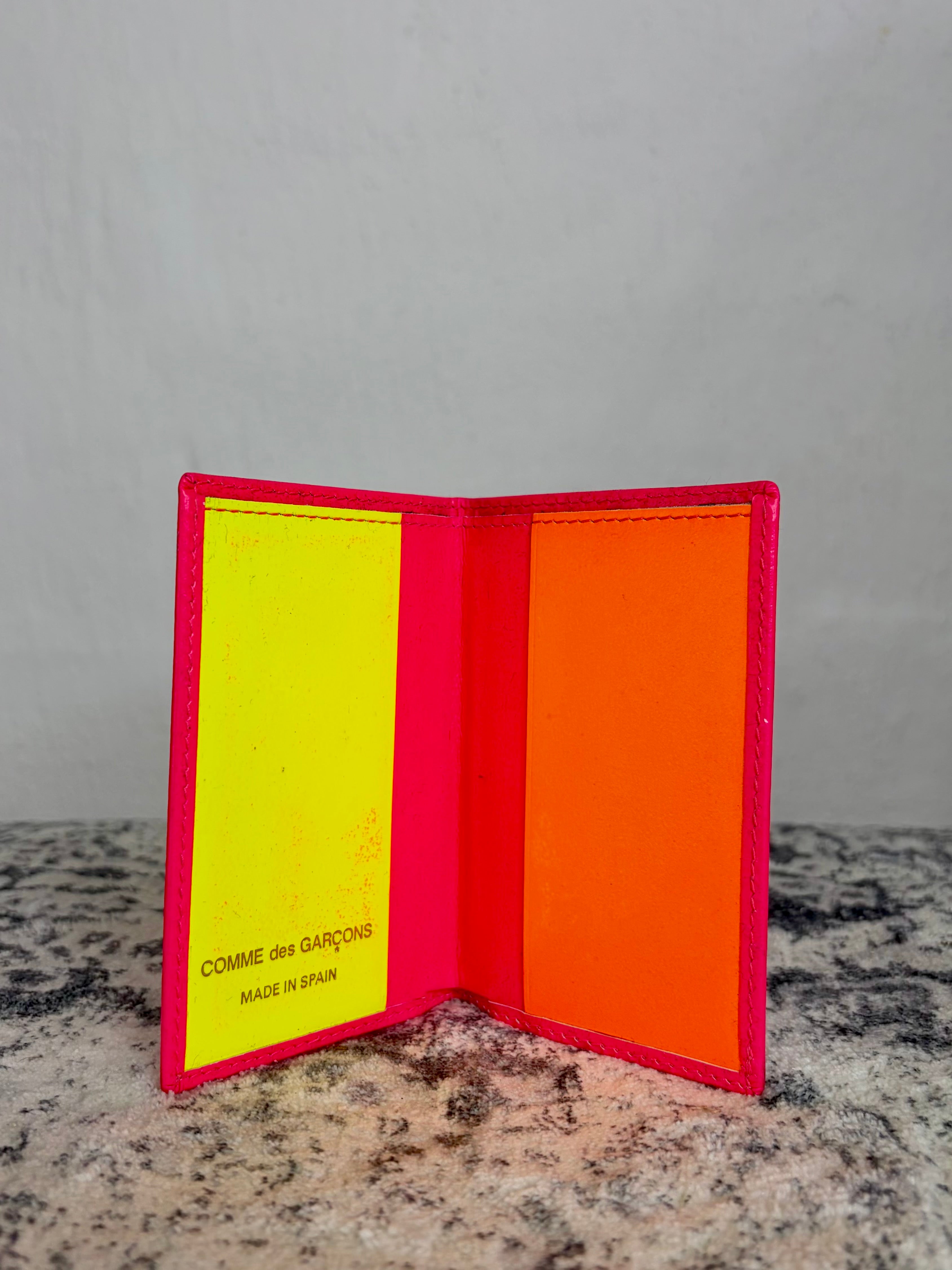 A bright pink leather bifold wallet standing open on a speckled stone surface, with a neon yellow card slot on the left and an orange interior panel on the right. The brand name 'COMME des GARÇONS' is printed in black on the yellow panel.