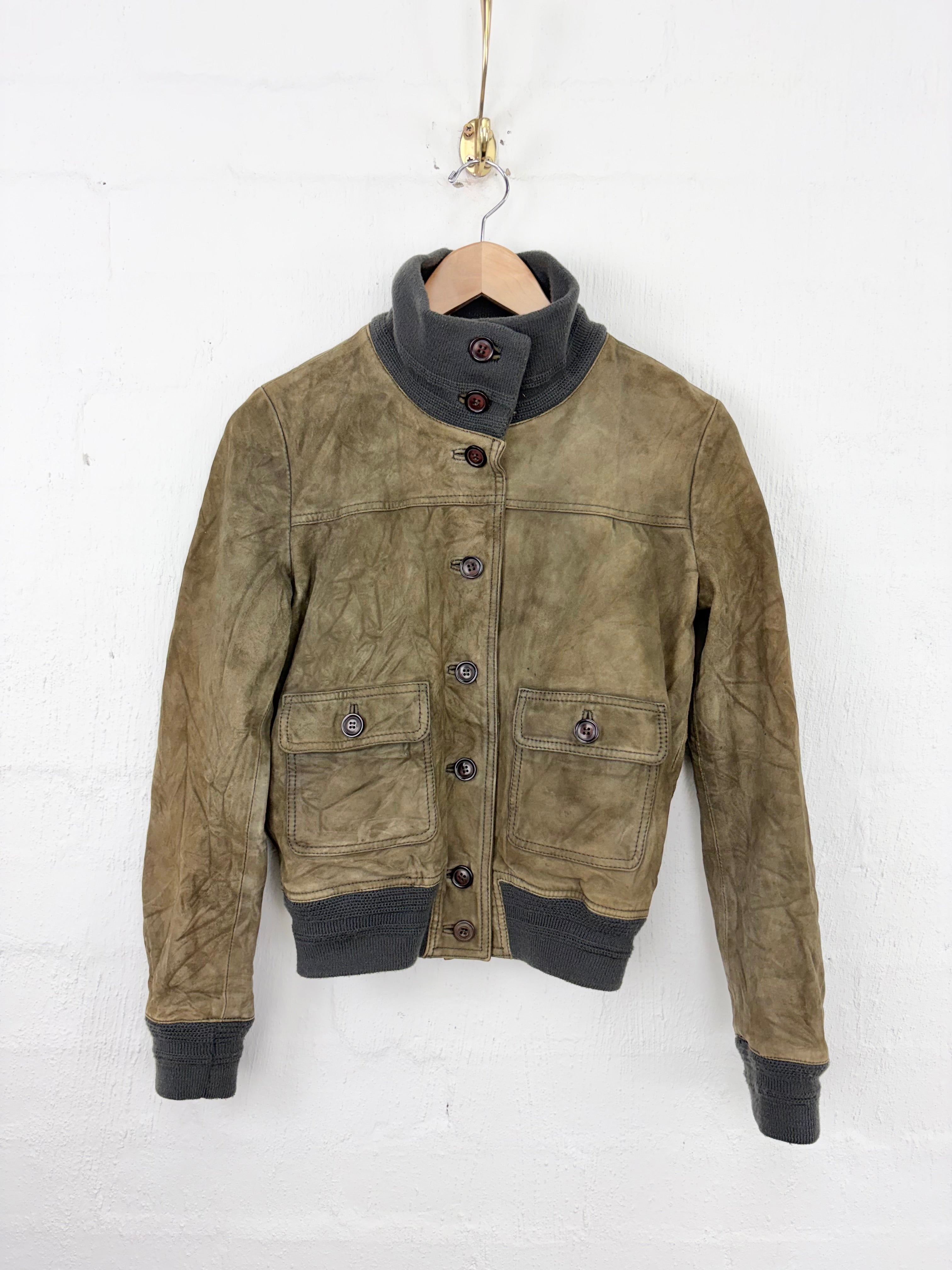 A vintage-style brown suede jacket with dark gray ribbed knit trim, hanging on a wooden hanger against a white brick wall.
